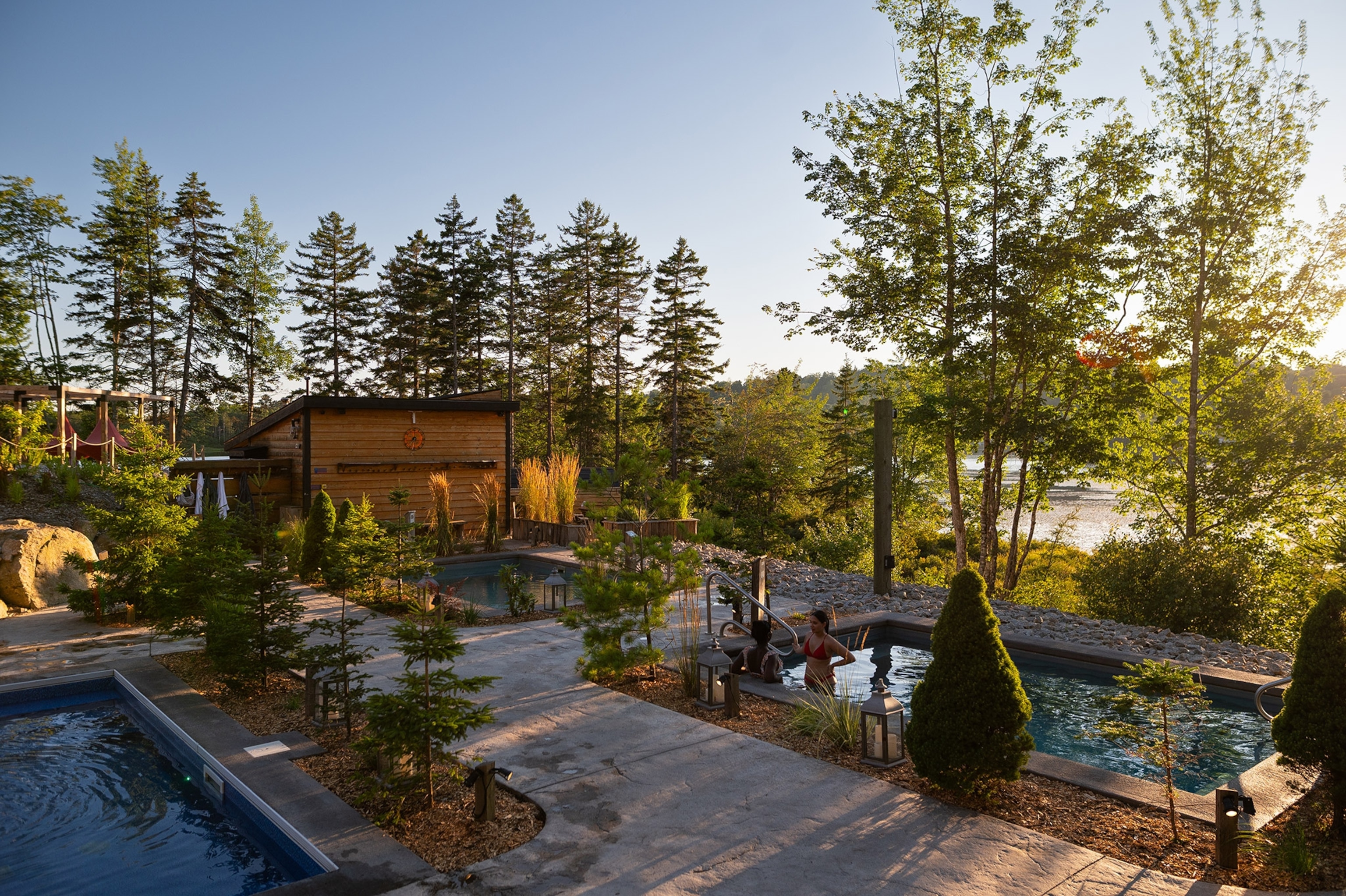 Outdoor pool deck with a small wooden shed, two small pools, surrounded by trees. In one pool, two women stand partially submerged