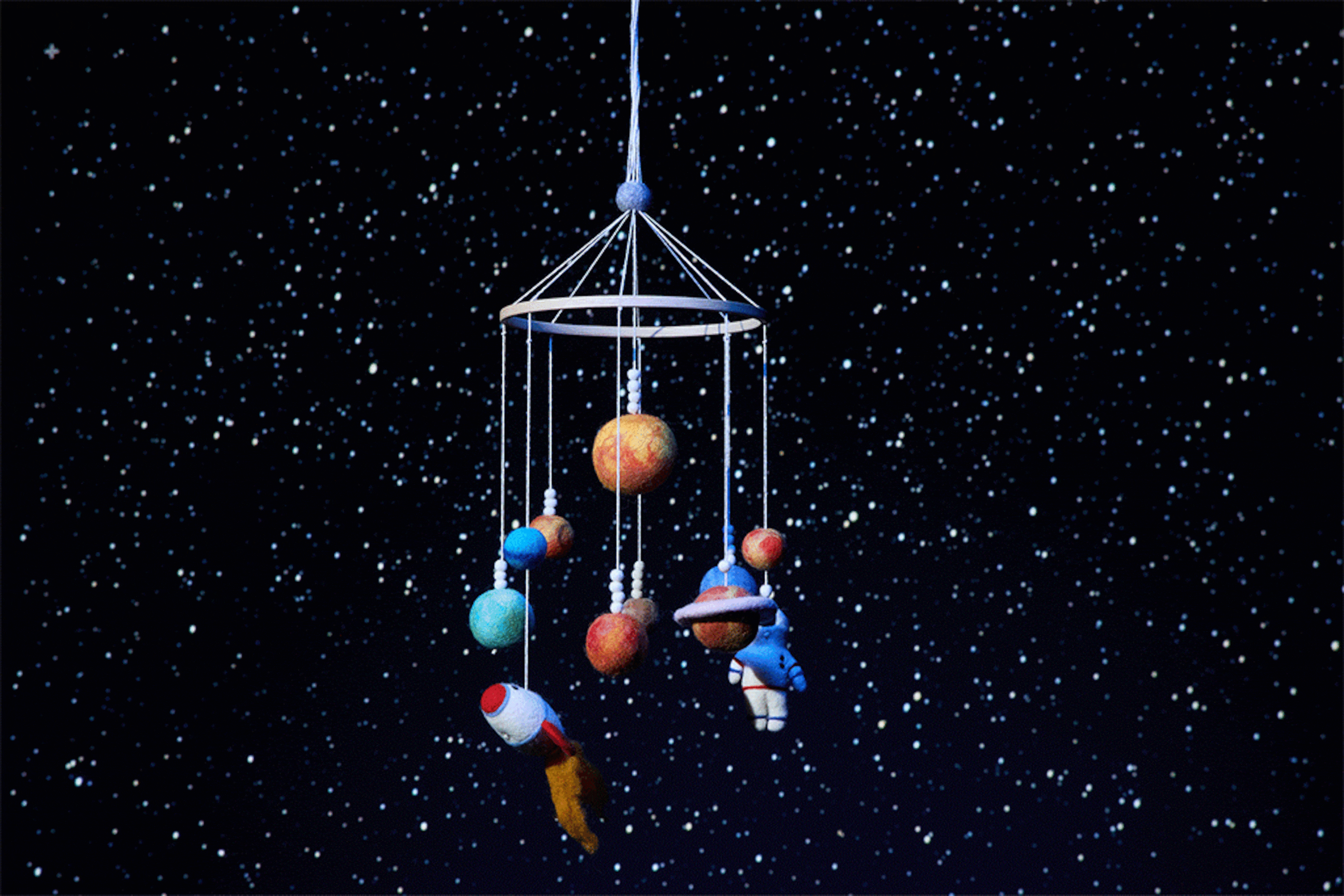 A space-themed mobile with an astronaut, rocket ship, and planets made of felted wool spins in front of a starry sky.