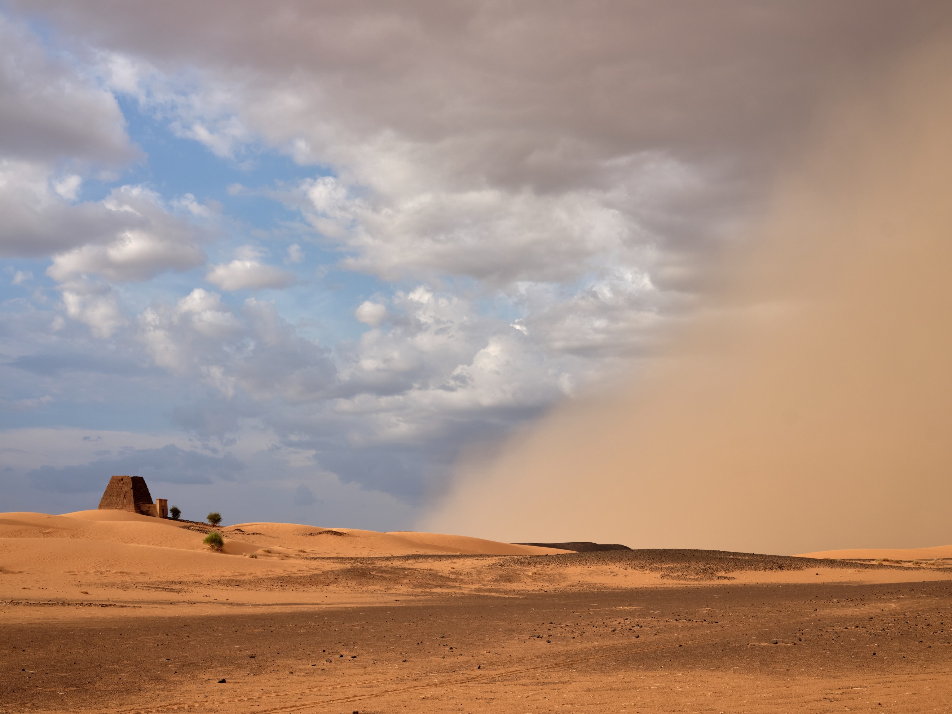 A trapezoidal-shaped pyramid is seen from afar, its size dwarfed by an approaching dust storm.