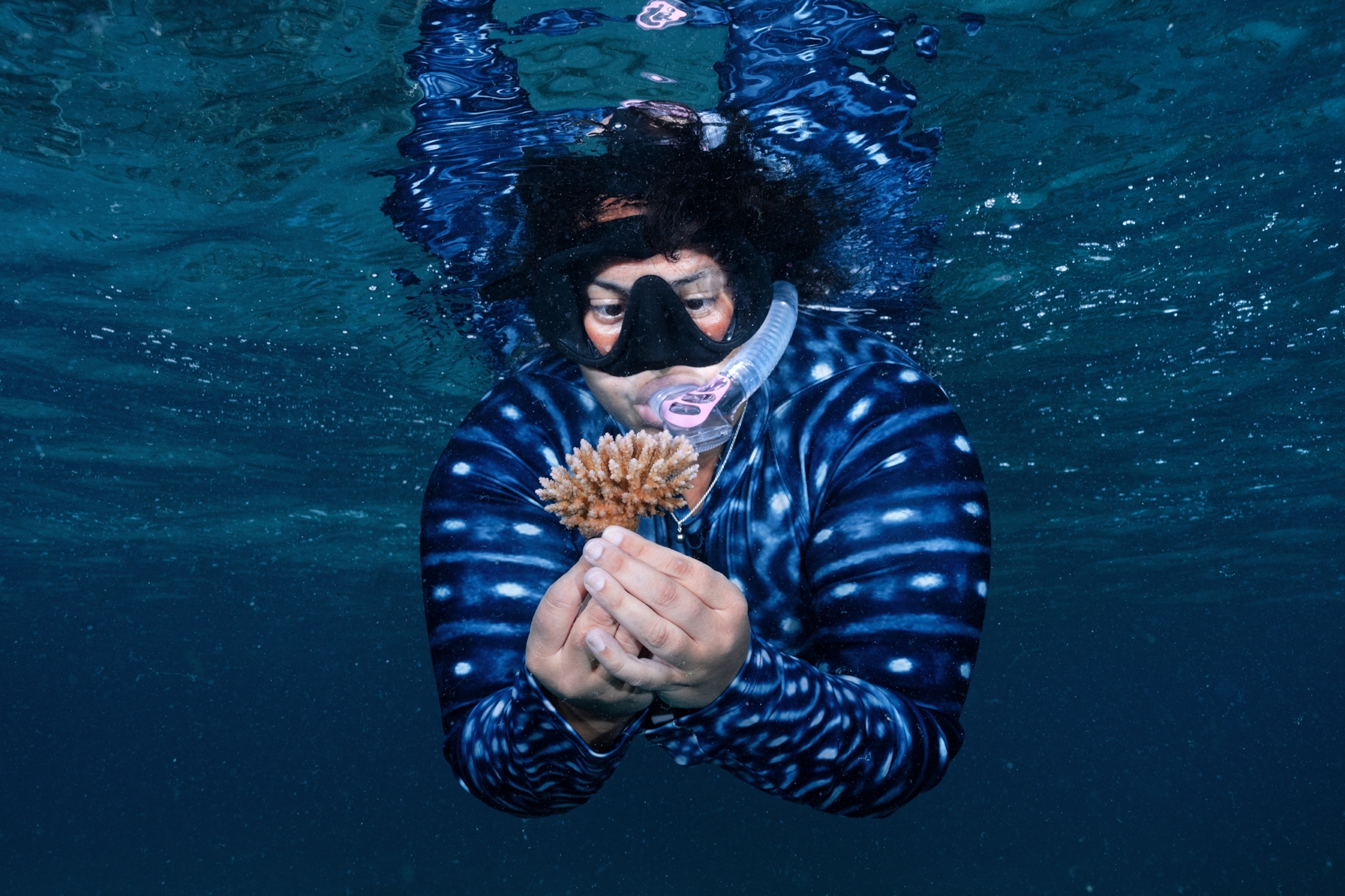 Underwater portrait of woman diver in polkadot suit and black mask looking at the coral bunch in her hands.
