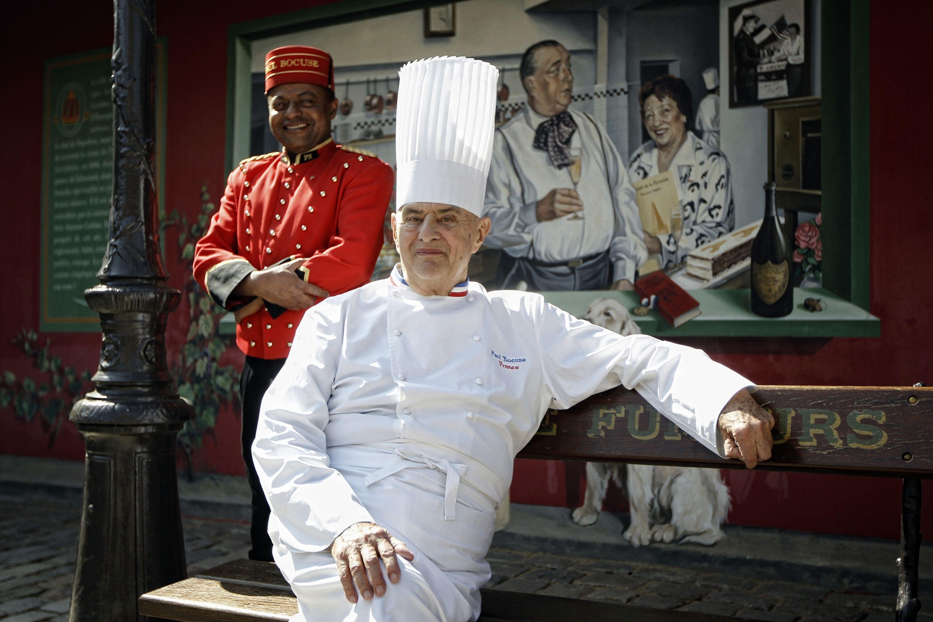 An elderly chef lounging on a park bench outside his restaurant wearing a chef's coat and tall hat.