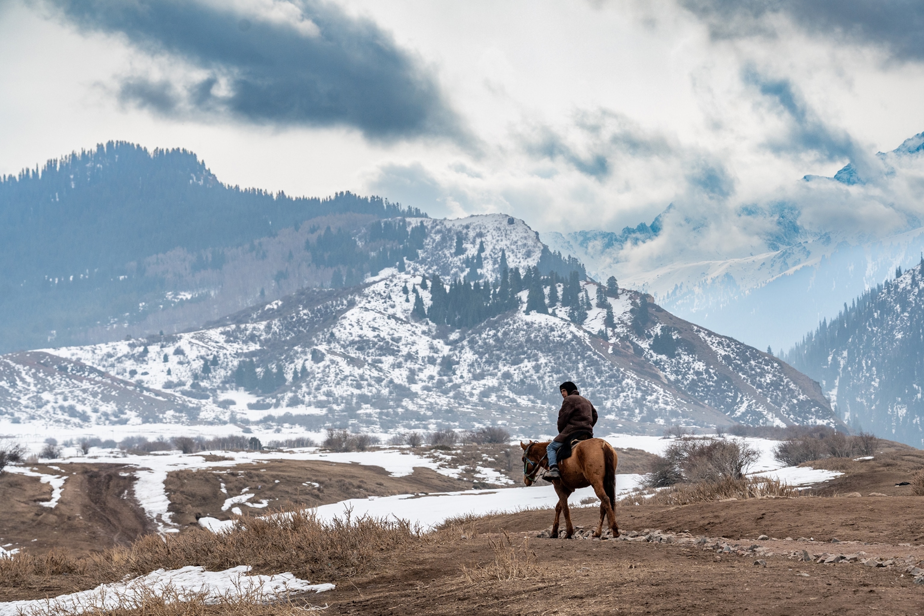 A lone man on a horse looking out across a high-altitude landscape with snowy mountains in the distance.