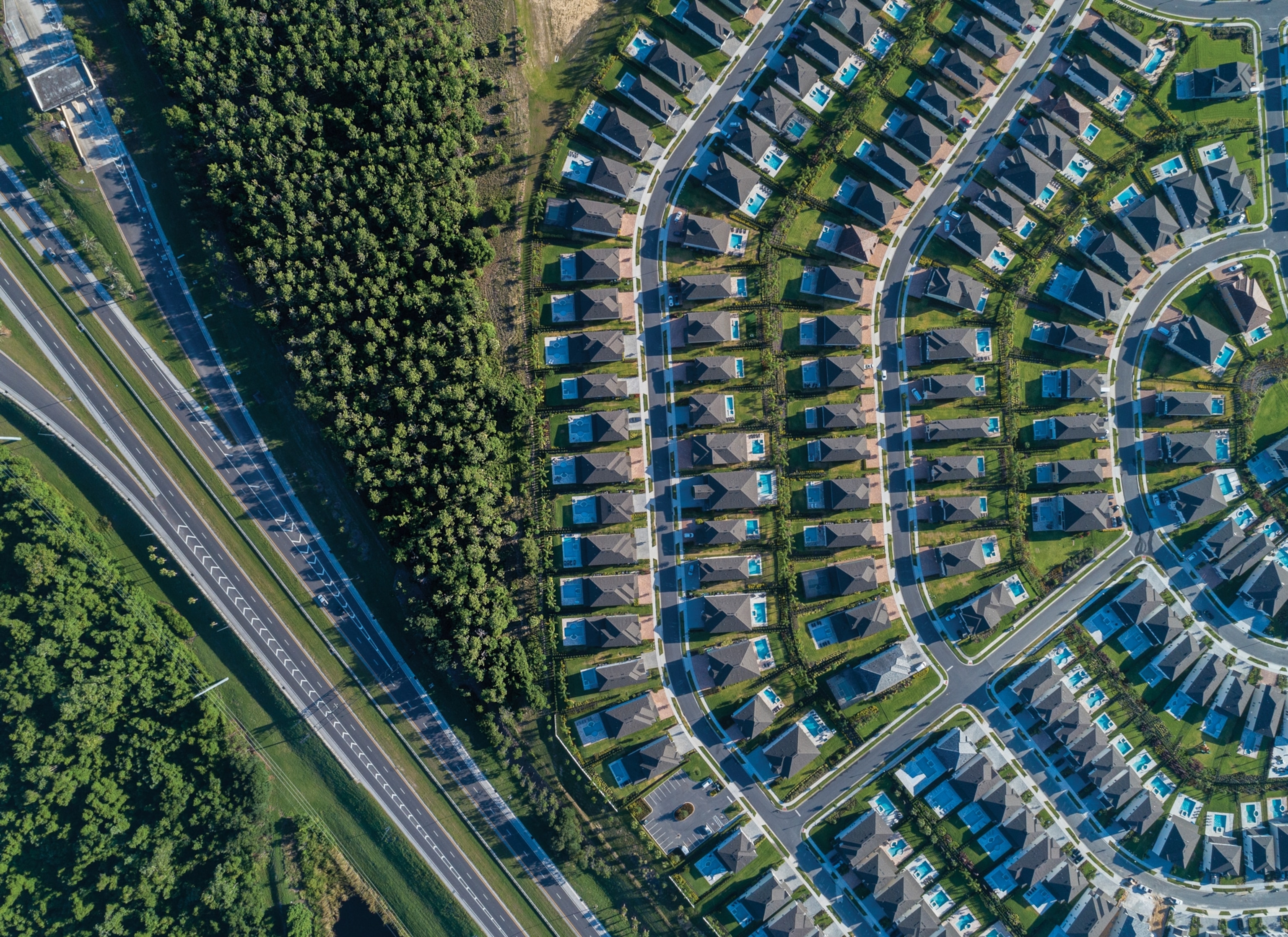 Aerial view of a suburban neighborhood with curved streets, uniform houses, and small pools. Dense forest borders the area on the left, next to a highway.