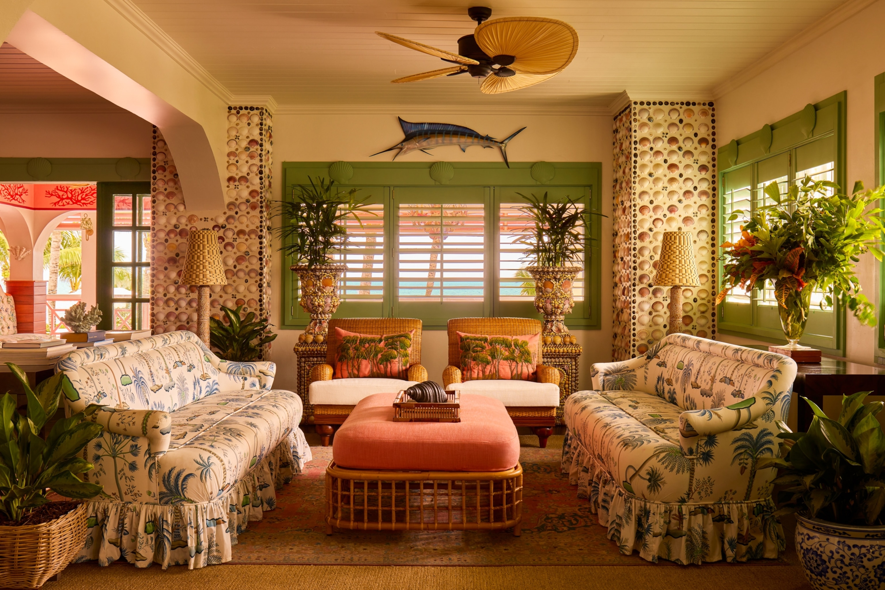 A colorful lounge with patterned sofas, wicker furniture, tropical plants, and green shutters facing the ocean.