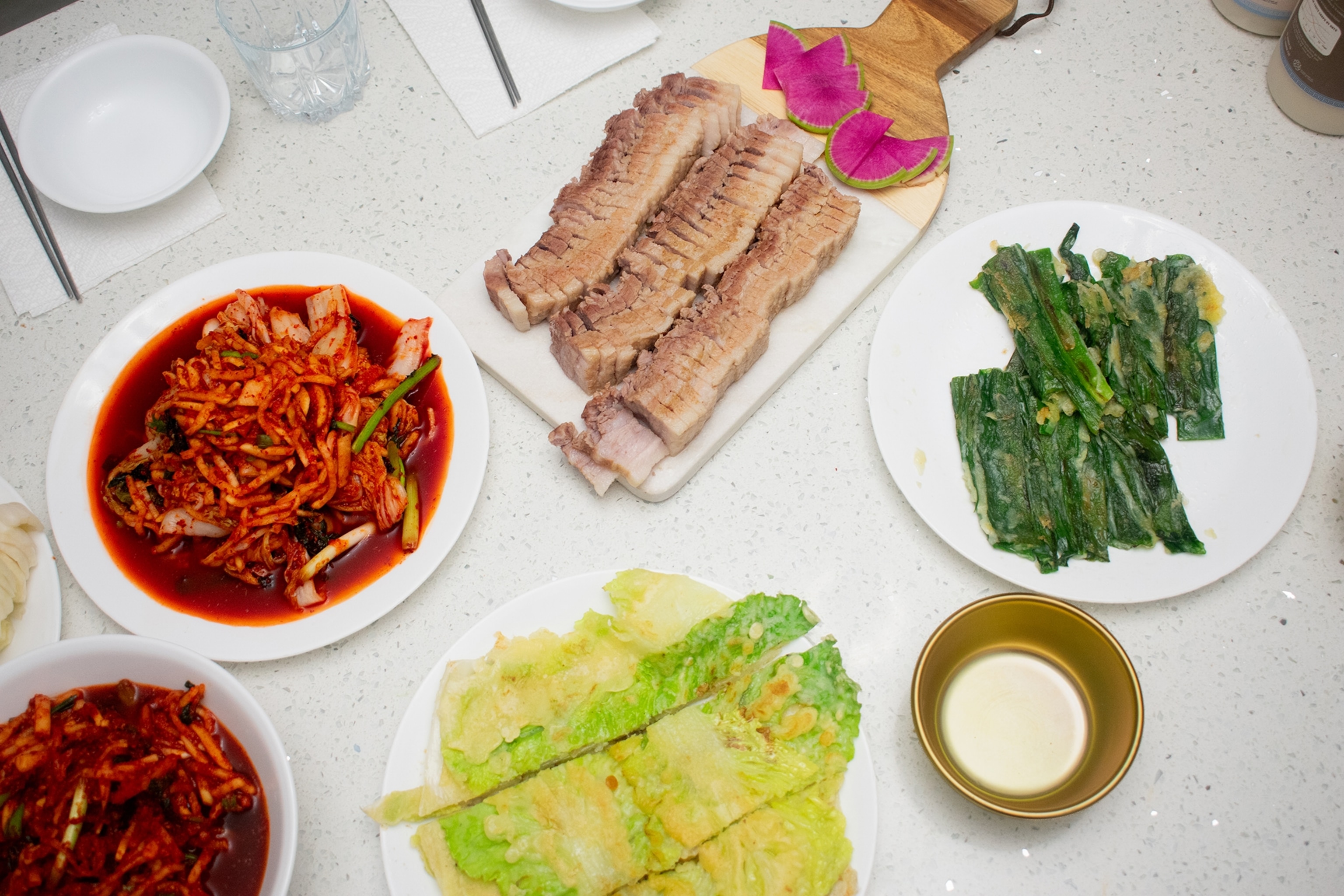 A spread of different dishes atop a marble kitchen island, including boiled pork belly, kimchi paste and cabbage pancakes.