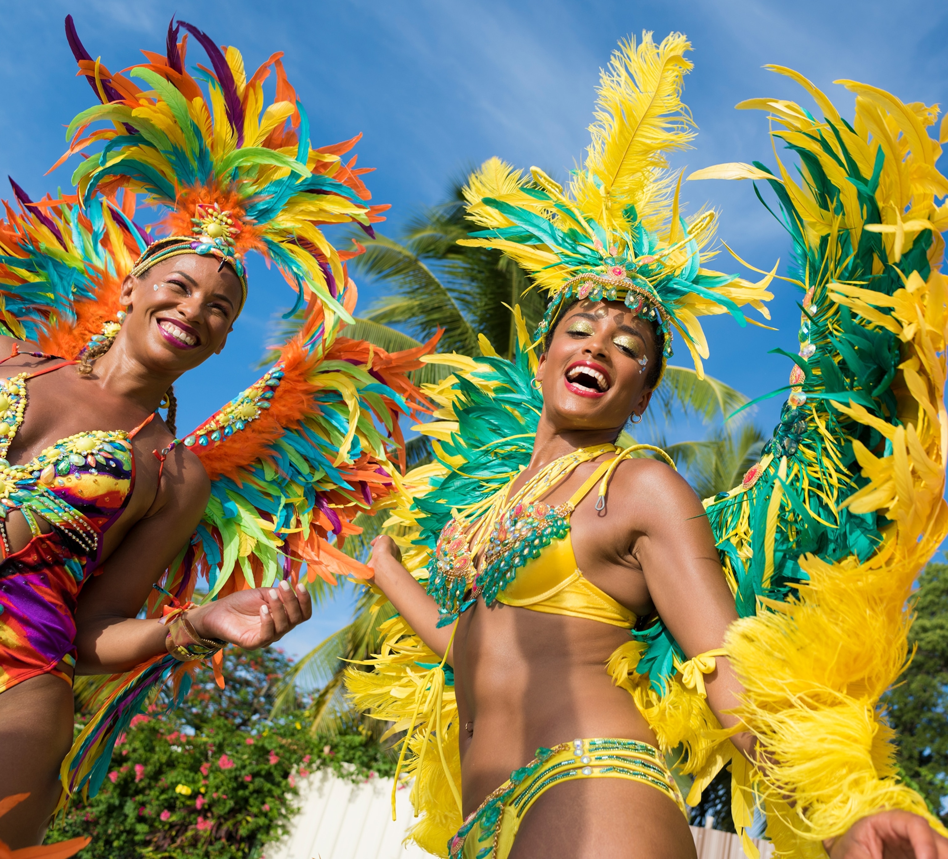 Two women in brighting colored large feathered costumes dance and smile looking down at the camera under a blue sky.