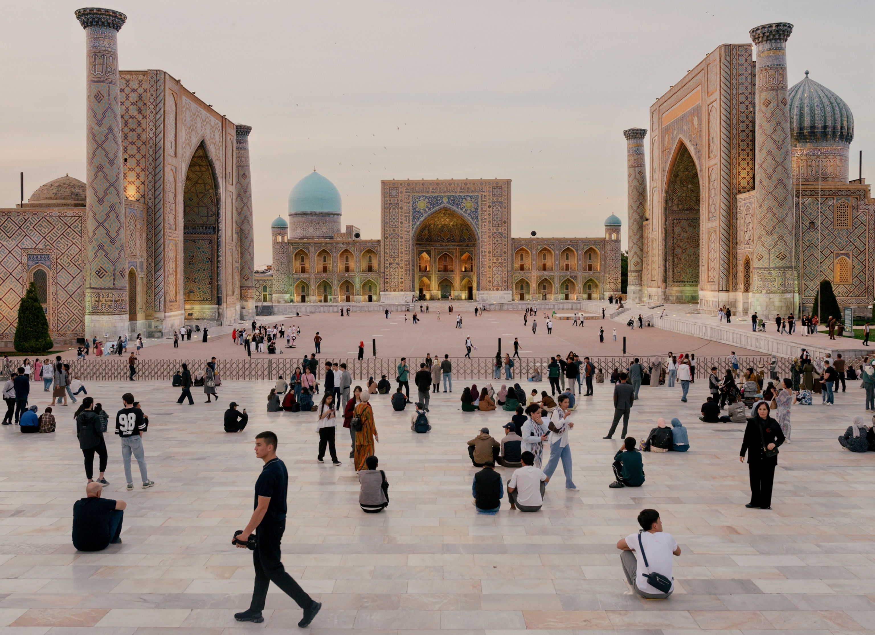 A wide landscape in a city, outdoor of a square tiled floor with arches and columns. Dozens of people walk about the square.