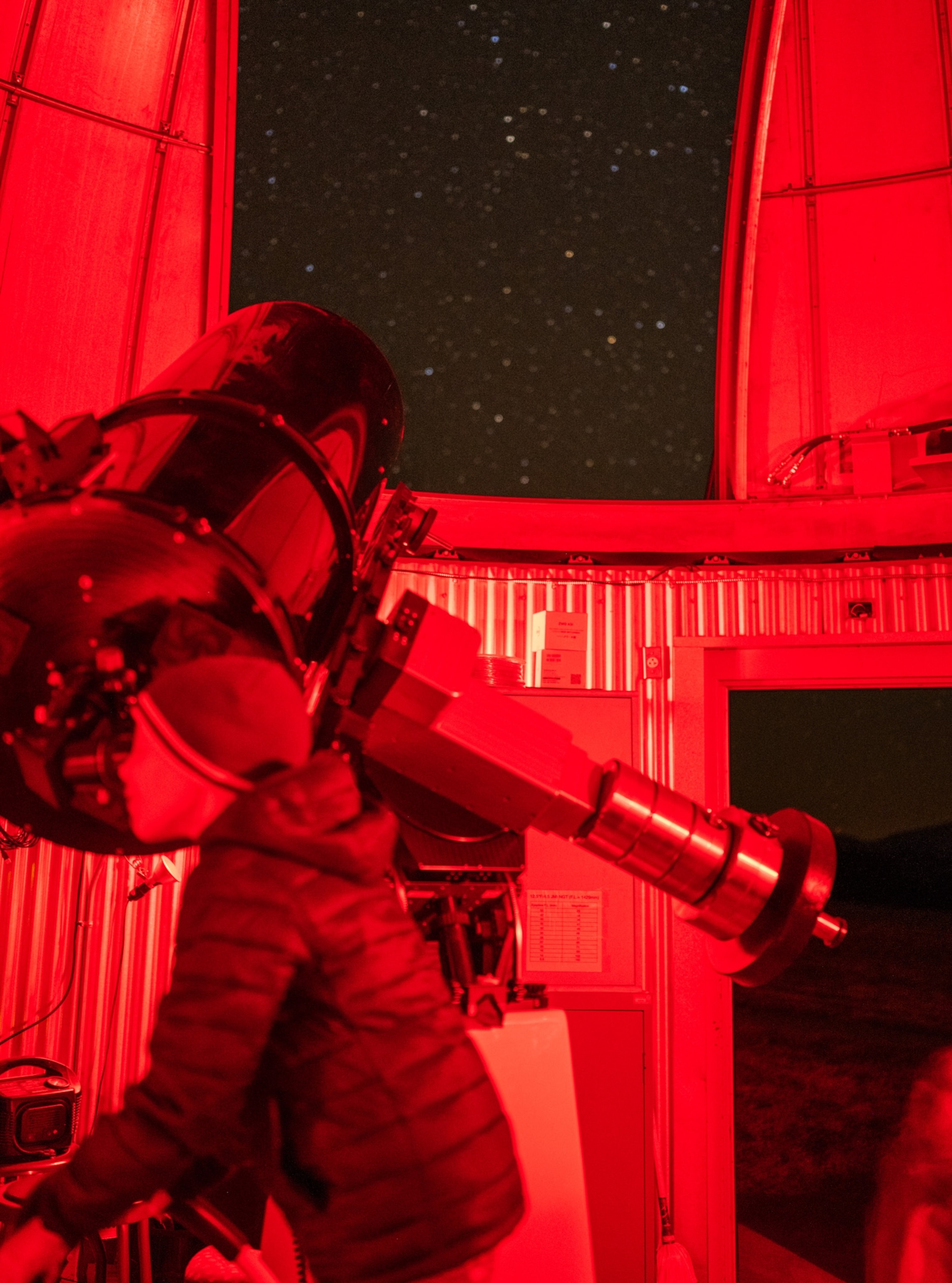A child wearing a hat and winter jacket looks towards a telescope. The area is red and the dark sky is filled with stars.