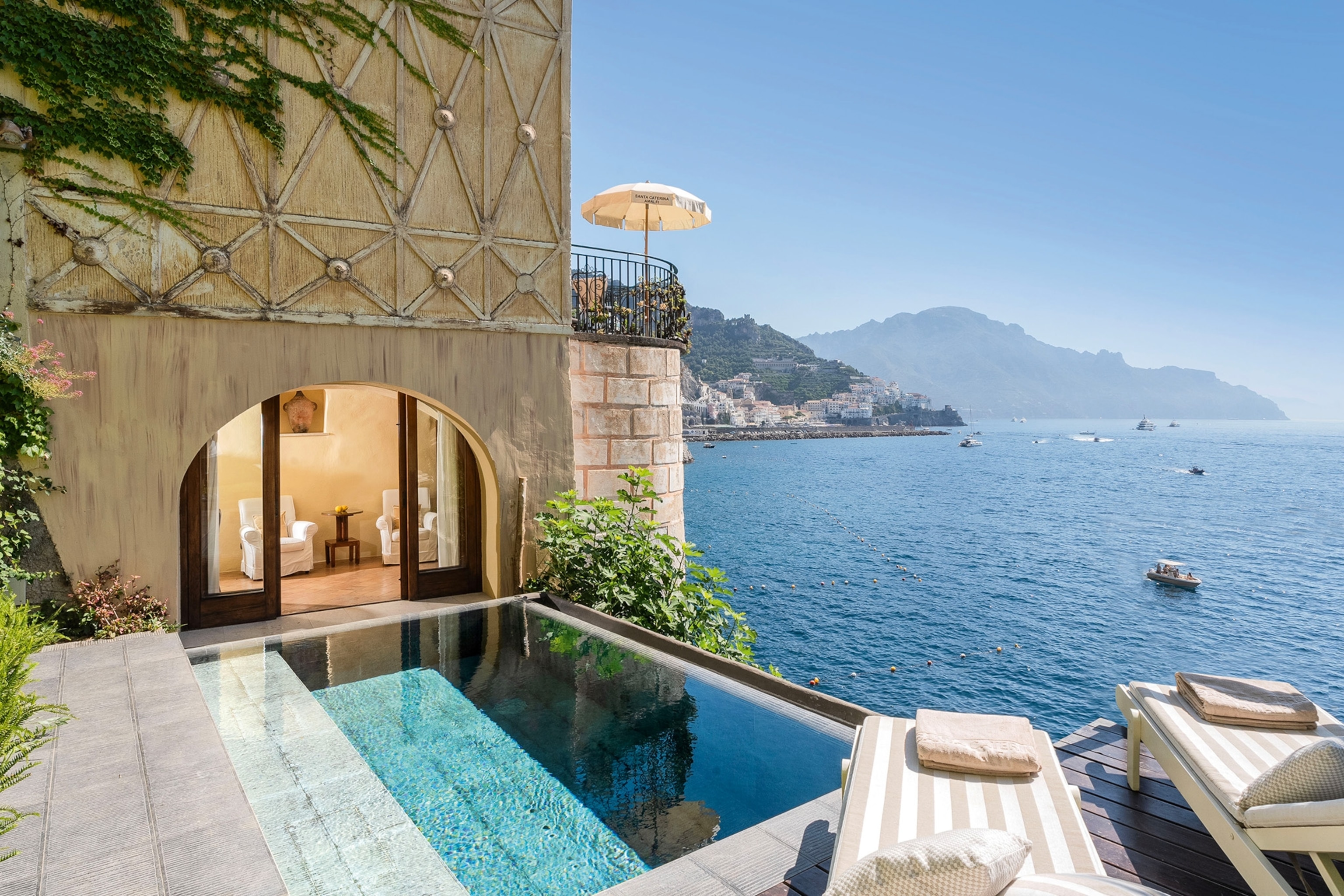 A pool with striped sun loungers sit on a cliff edge overlooking the sea.