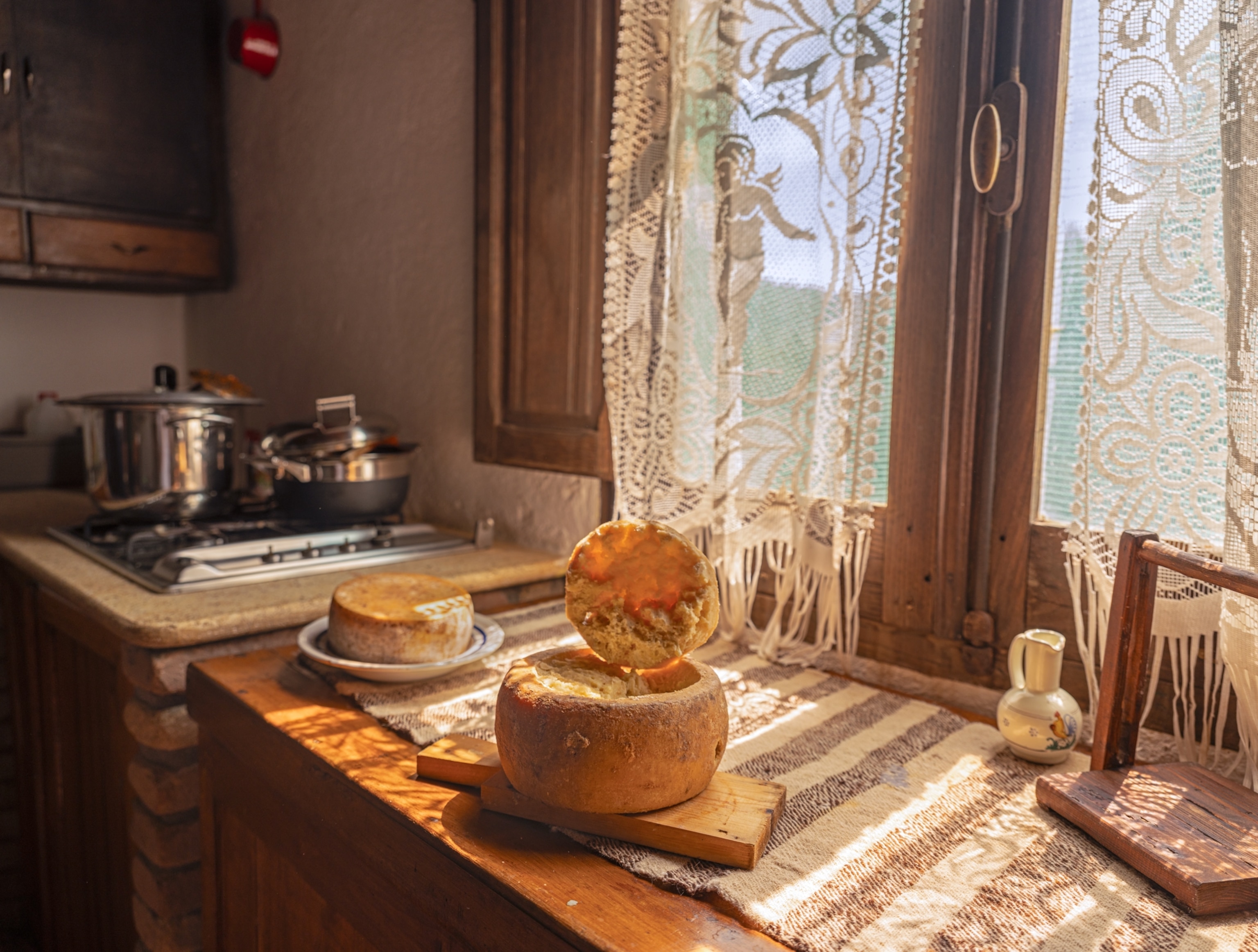Country kitchen with pots and pens and two cheese wheels on the table by the window in the sunlight penetrated through lace curtains.
