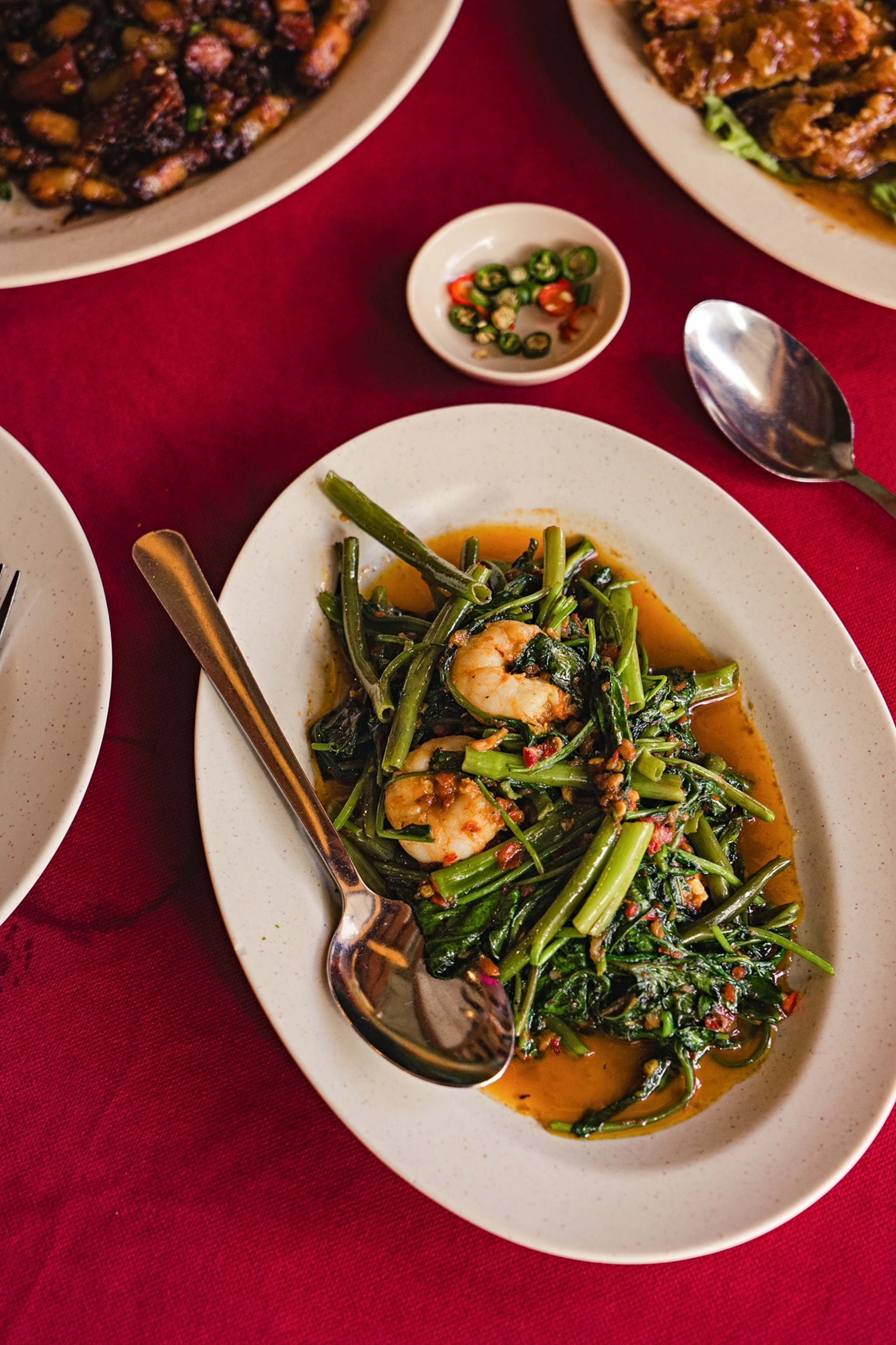 A dish with shrimp and greens against a red table cloth. Other plates of food surround the edges of the frame.