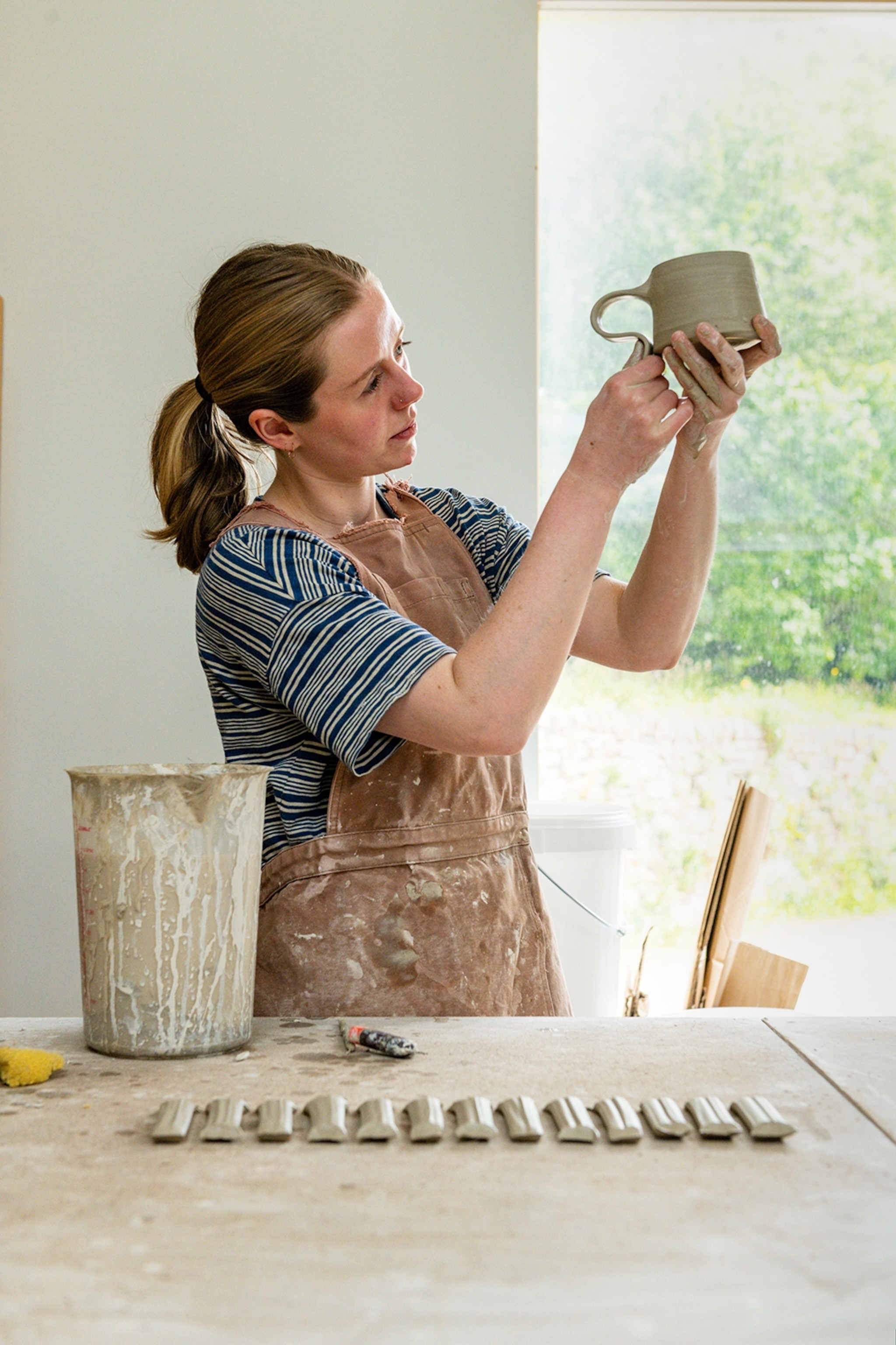 A young woman shaping a handle and attaching it to a clay mug in a studio.
