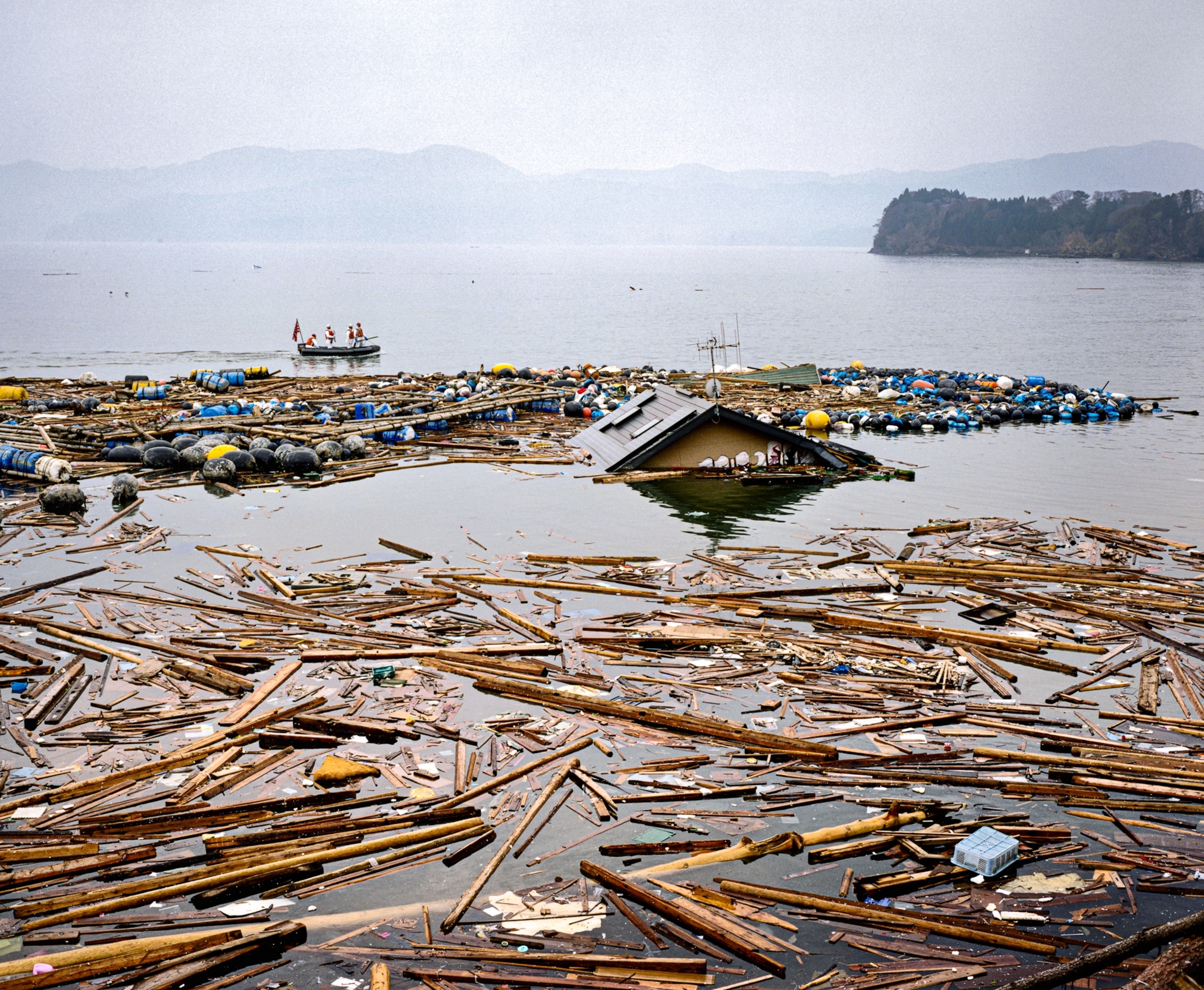 People on the boat going around floating debris and house's roof in the middle of it.