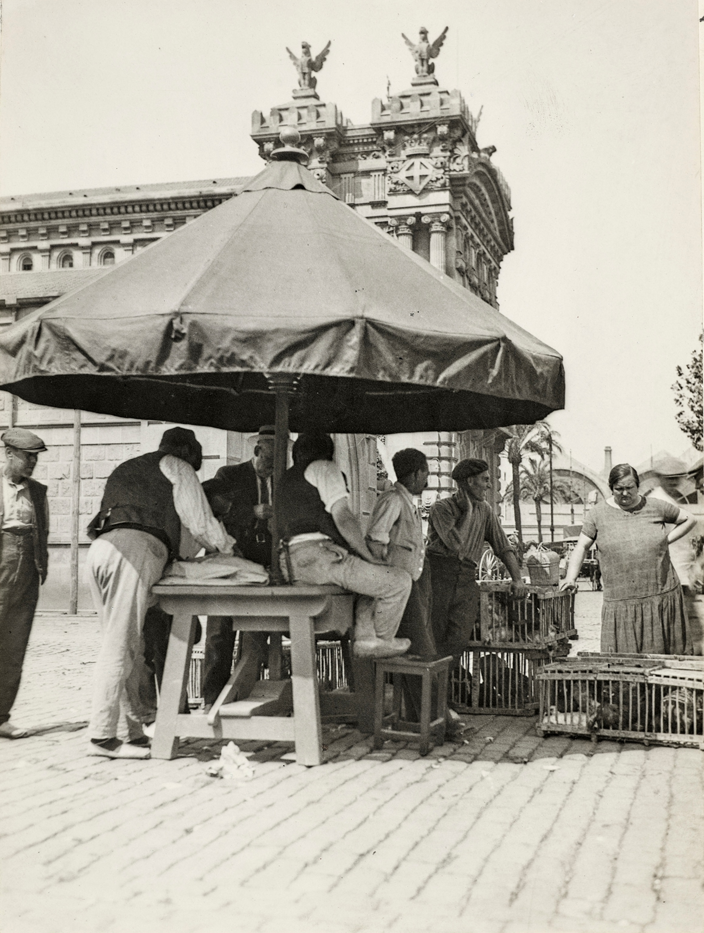 A view of people at the waterfront with crates of chickens that just arrived in the early morning on a ship from Majorca (Mallorca) and an aduana (custom house) in the background.