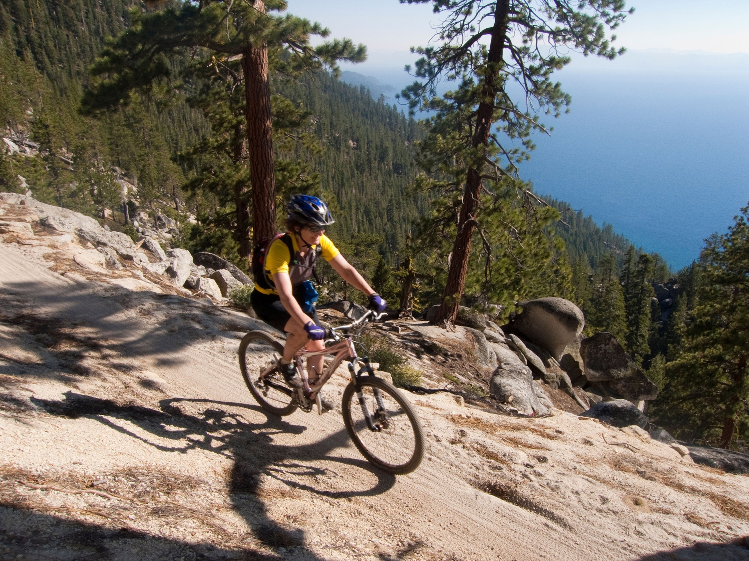 A cyclist in a yellow shirt and helmet rides a bike along a narrow trail on a high, tree-lined ridge overlooking a blue lake.