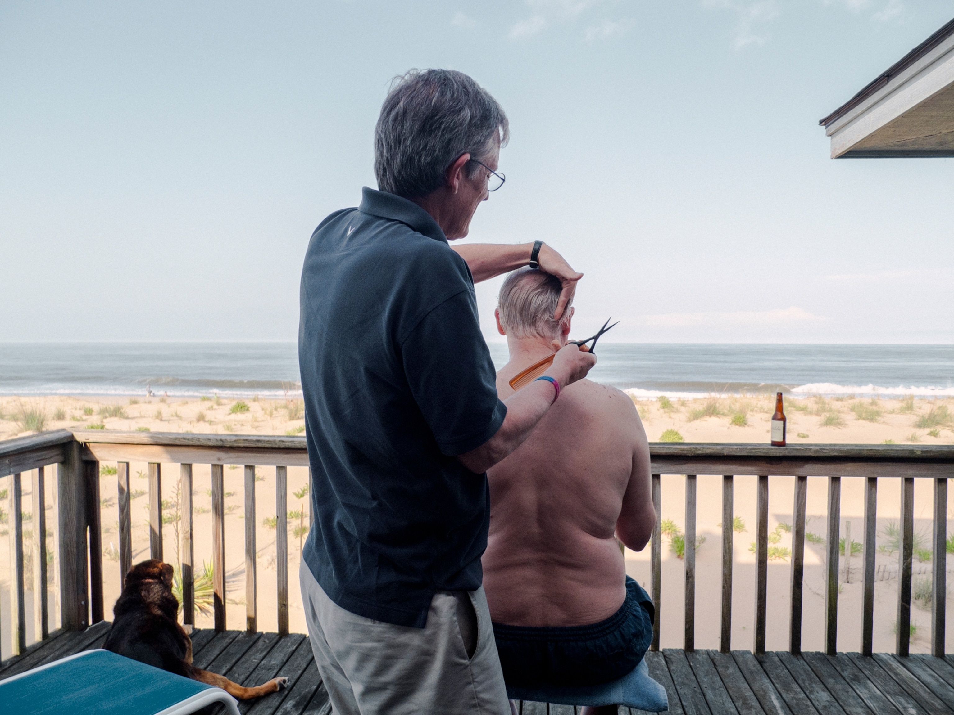 a man in a navy polo giving a shirtless man a haircut on the deck of a beach house facing the ocean
