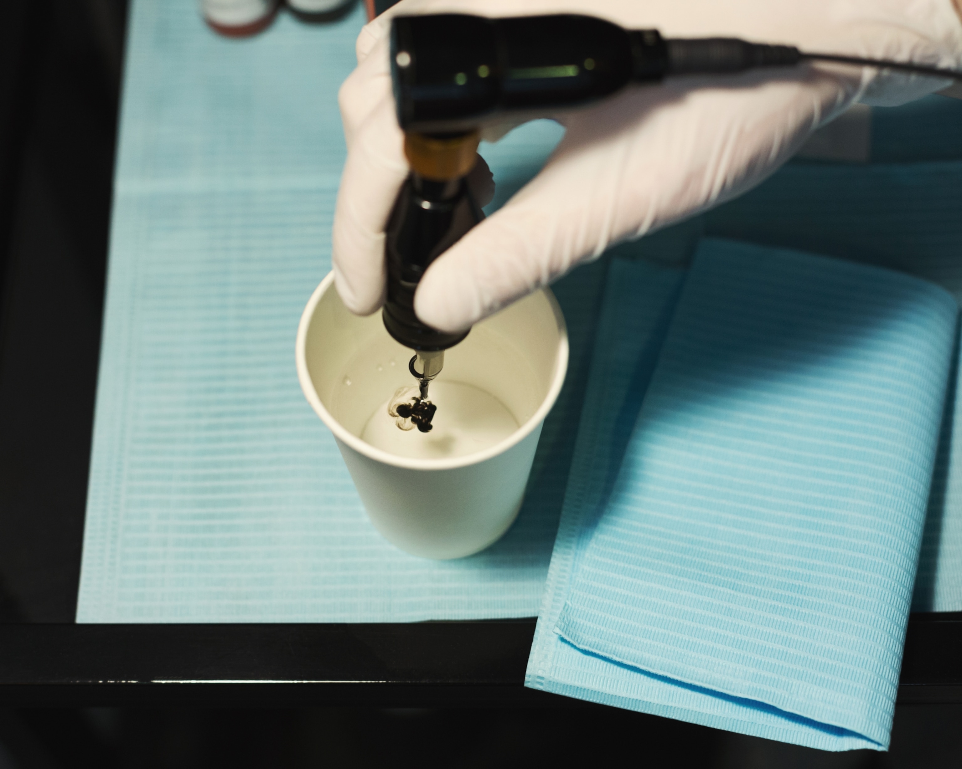A gloved hand hovers a tattoo machine over a white paper cup, preparing ink. The setup is on a blue medical sheet