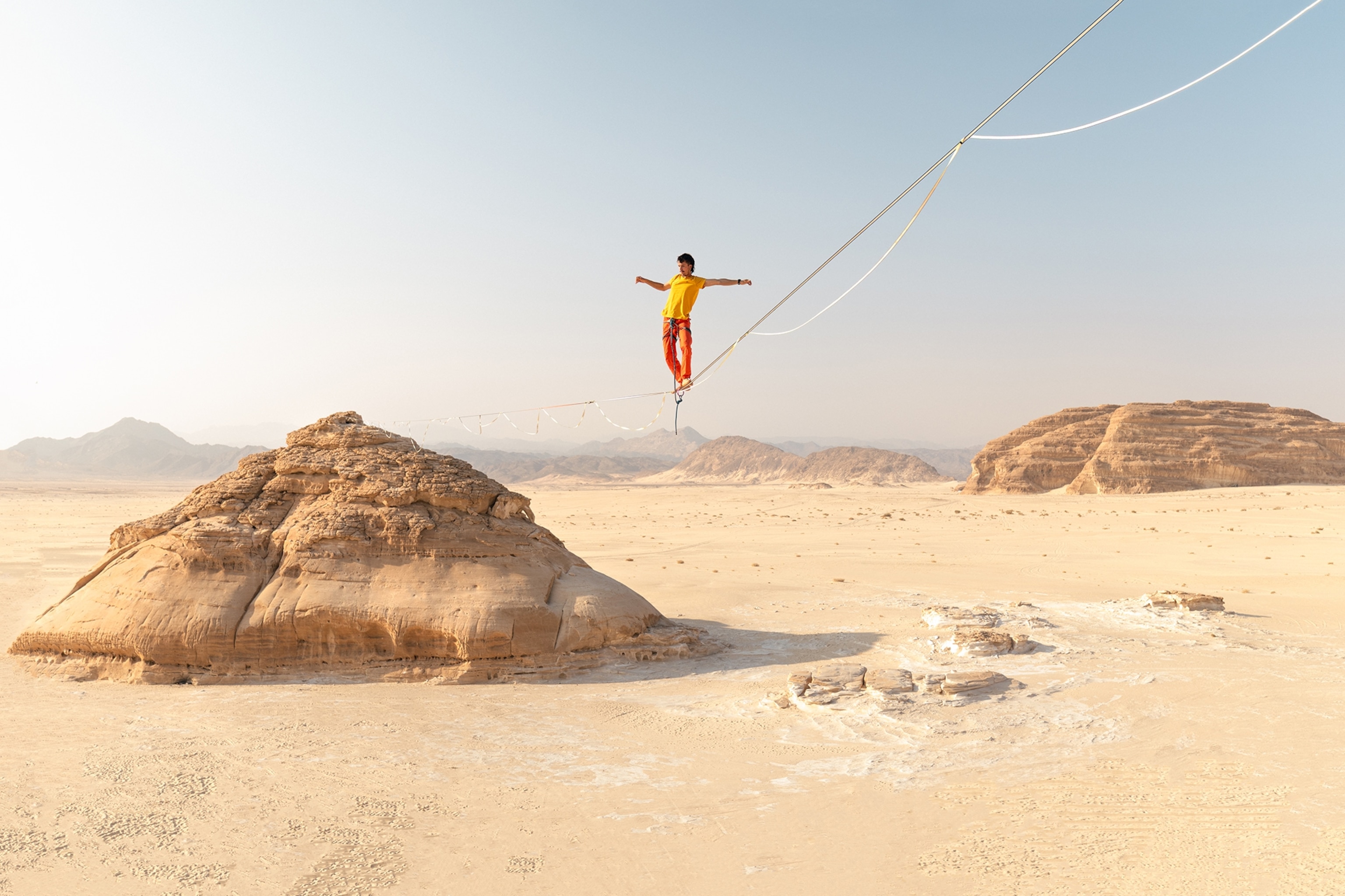 A tight rope in a desert with a young man balancing across it.