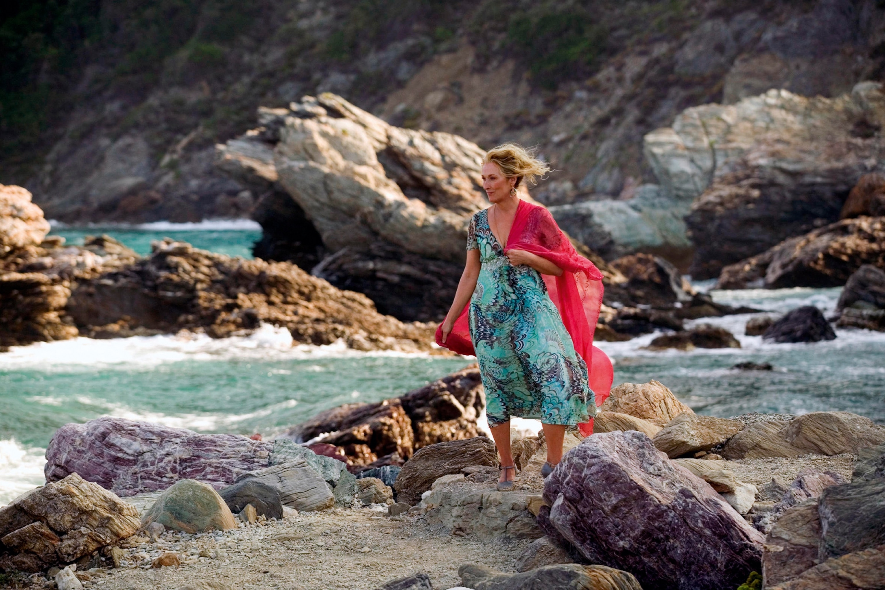 Meryl Streep in character on a rocky path with the water crashing into rocks behind.
