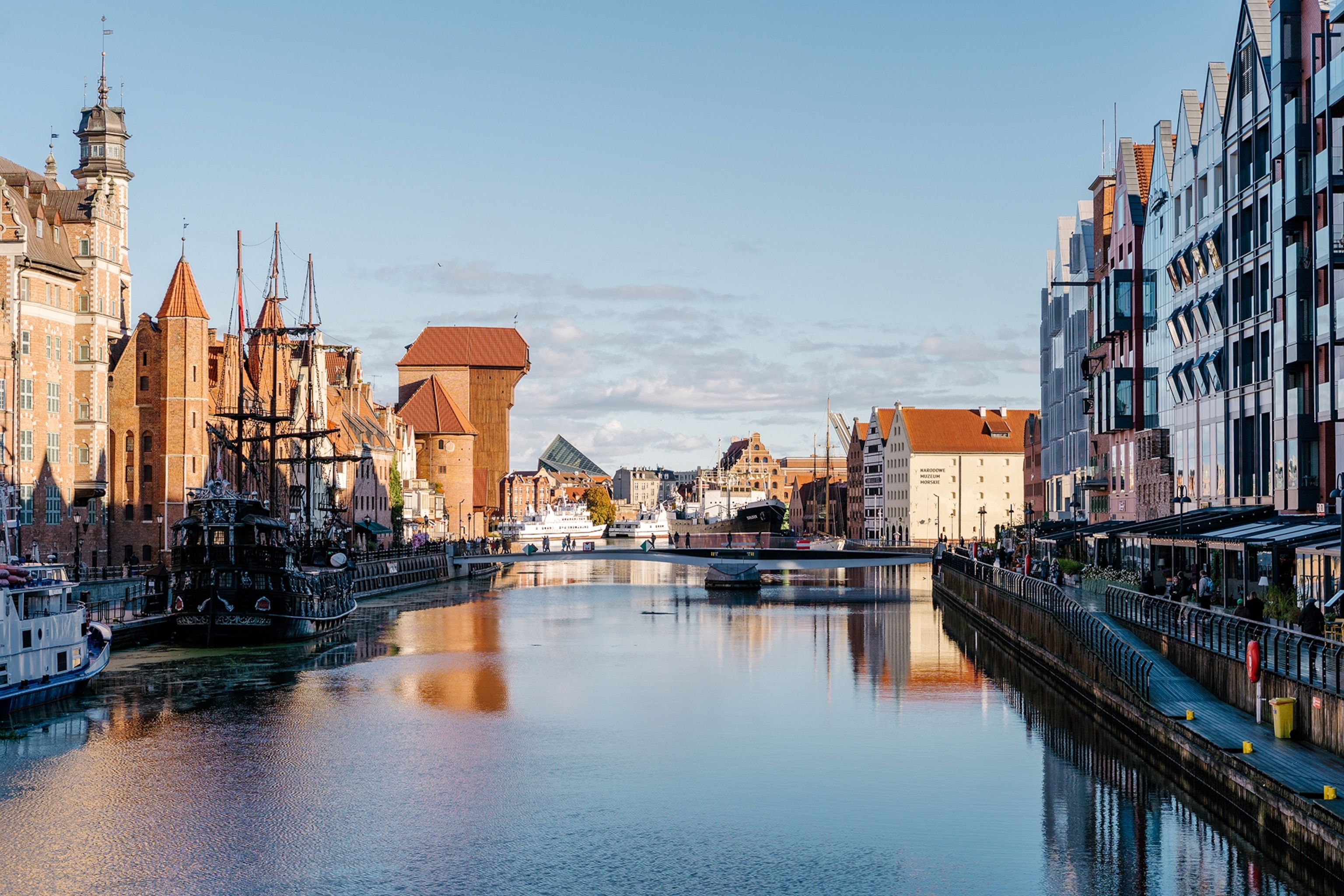 A wide, urban river running past gabled houses with both old and new boats anchored on either side.