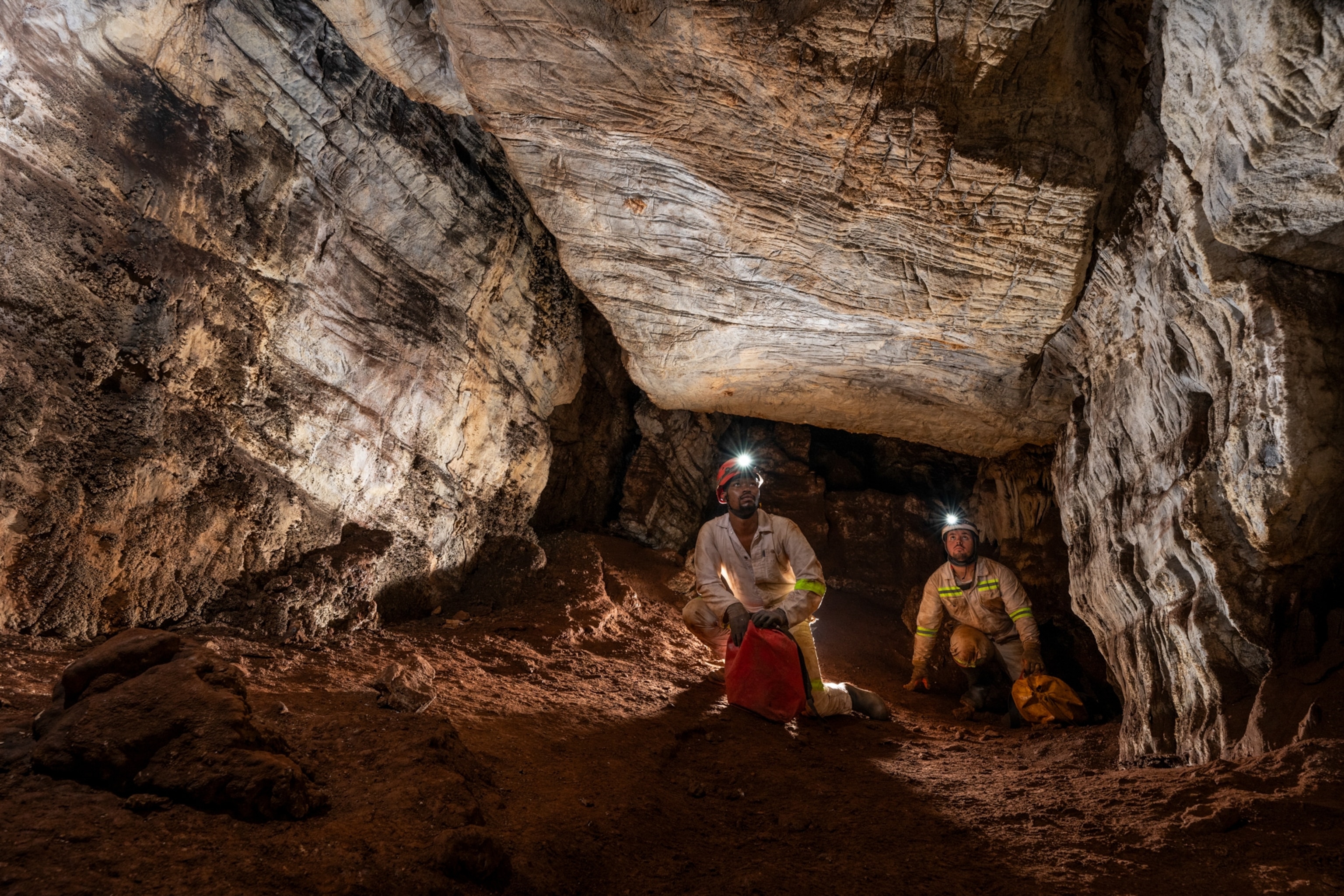 Two cavers with headlamps exploring a cave, surrounded by rocky walls and a dirt floor.
