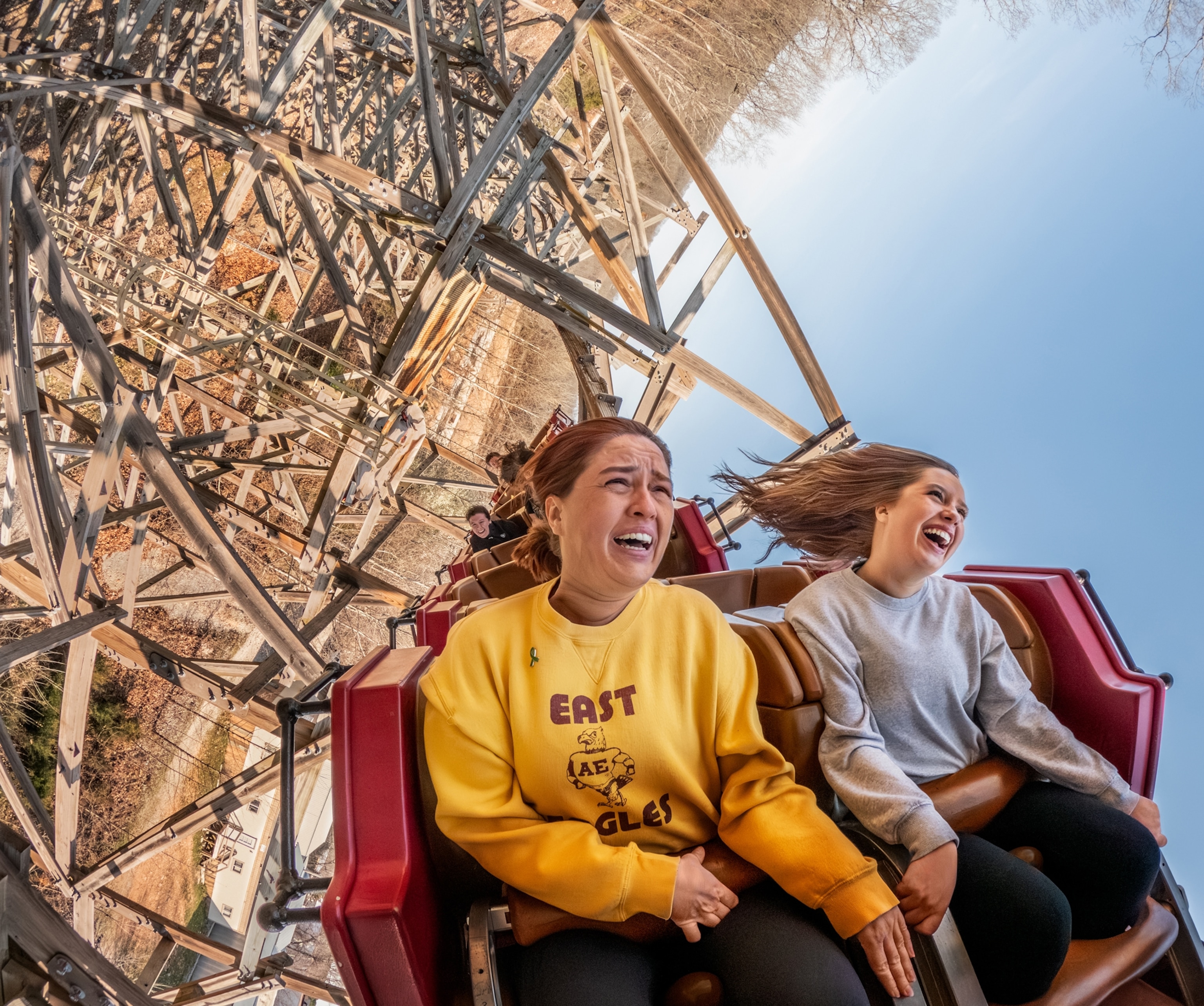 A mother and daughter sit in a red bench seat on a rollercoaster. The rollercoaster is in the middle of a loop, and the wooden structure can be seen behind the two people.