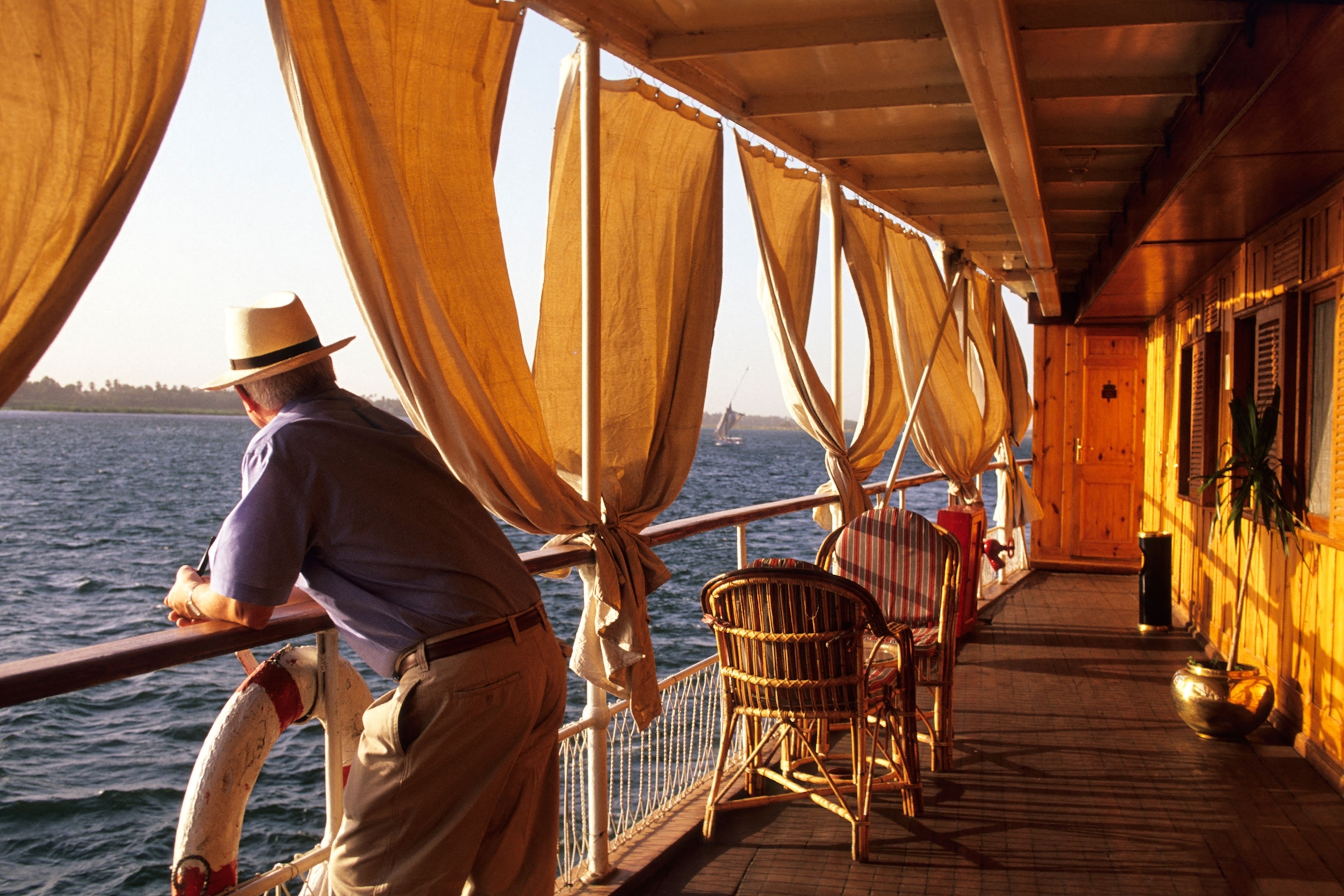 A man with fedora leaning on the railing of a river cruise ship, looking into the distance at sunset.