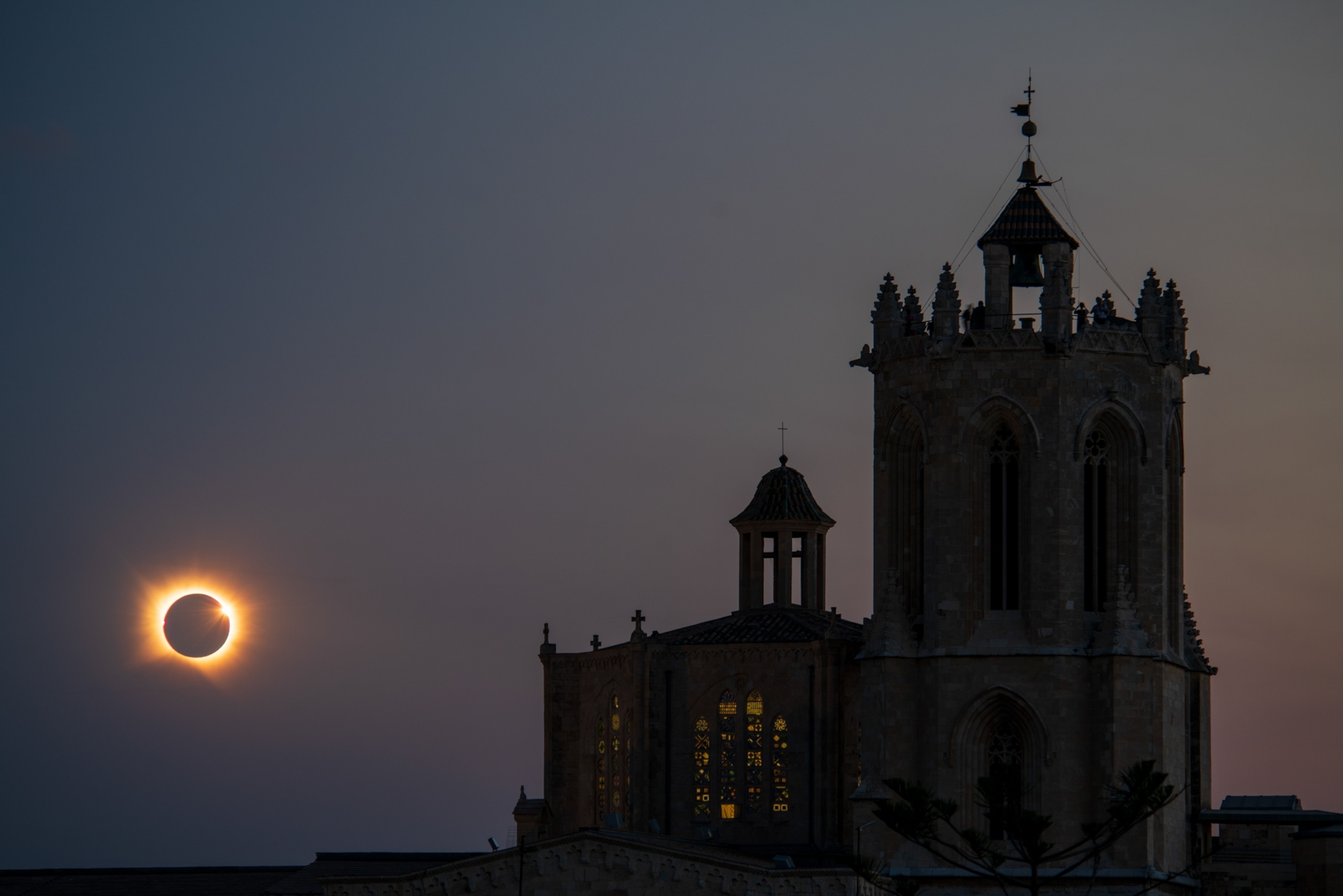 A glowing solar eclipse hangs beside a dark, ornate church tower, with warm light shining through stained-glass windows in a quiet twilight scene.