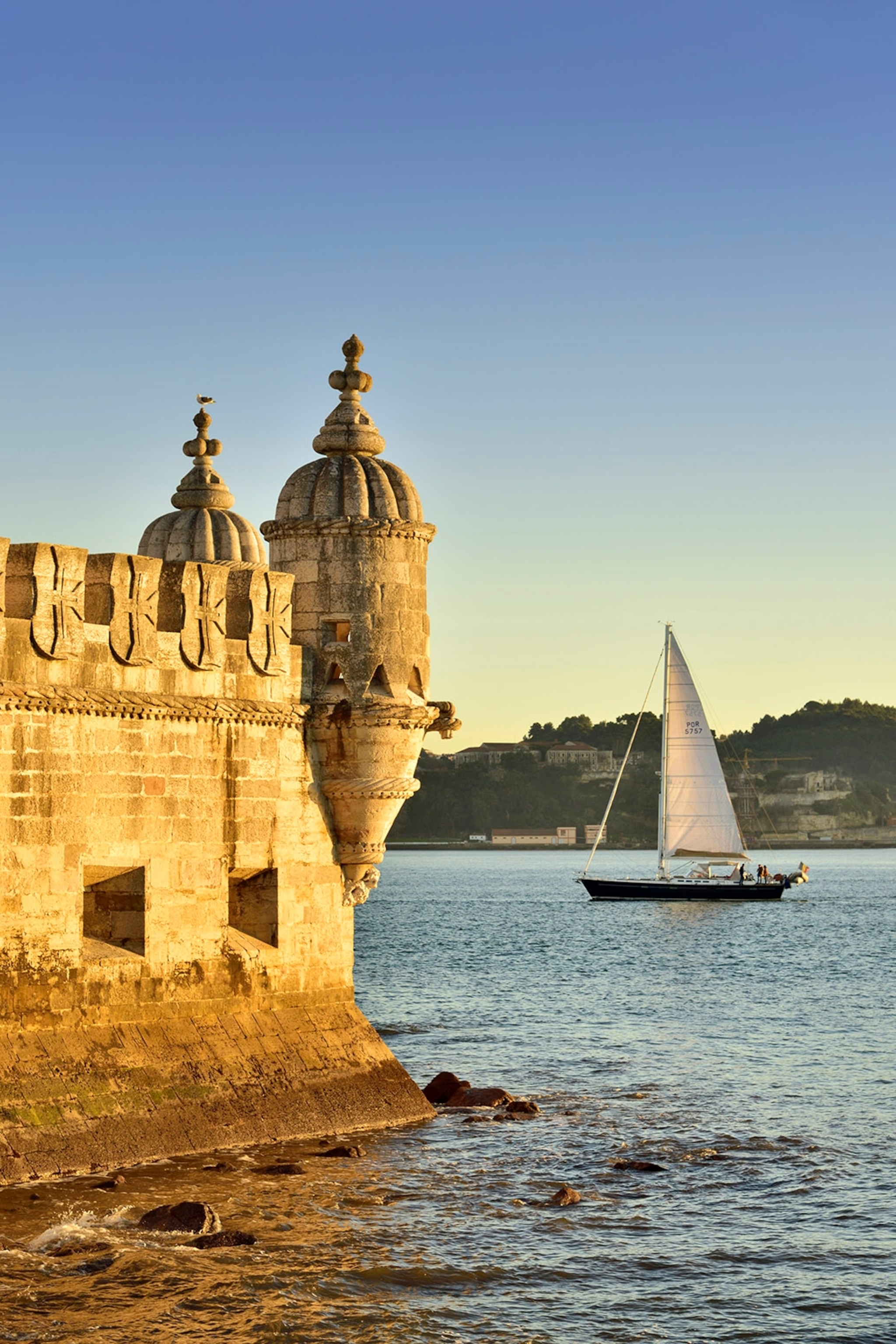 A sailing boat passing a stone fort at dusk.