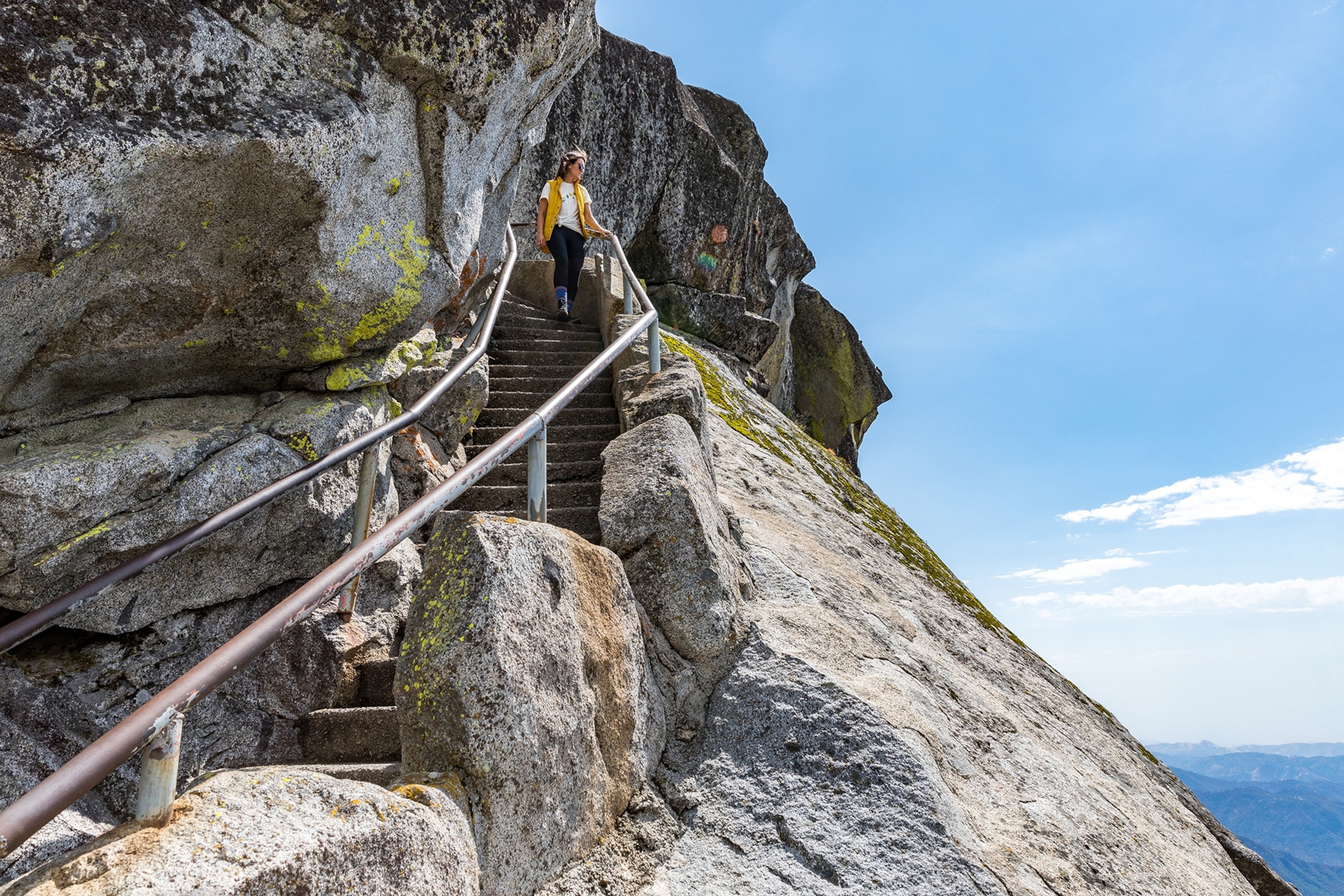 Woman walking down stairs embedded into a mountain holding onto a rail, while looking out into the distance