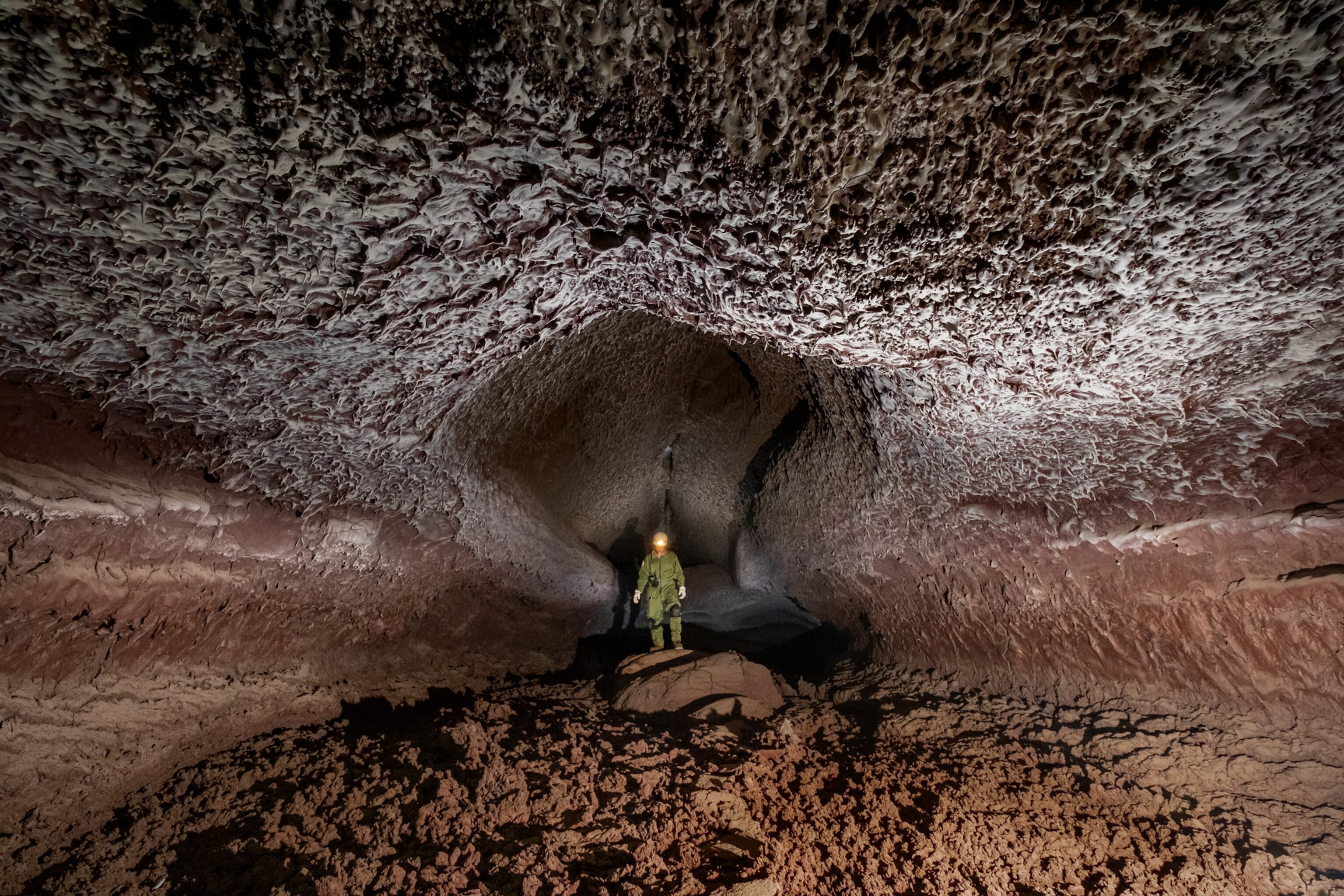 A scientist standing at the entrance to a large lava tube chamber, the walls a hue of reddish-brown, while the ceiling is a deeply textured white.