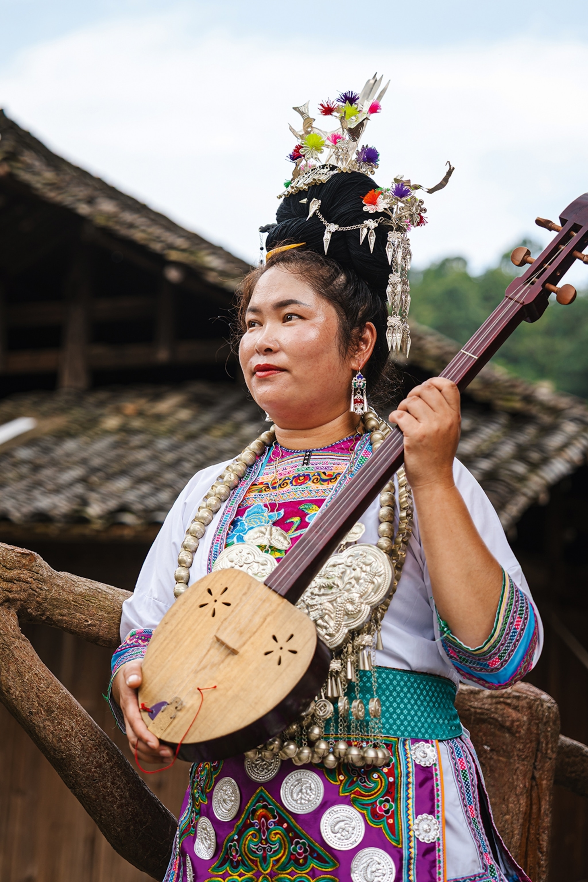 A Chinese woman in traditional dress and head piece holding a banjo-like guitar and standing in a wooden house villages in the mountains.