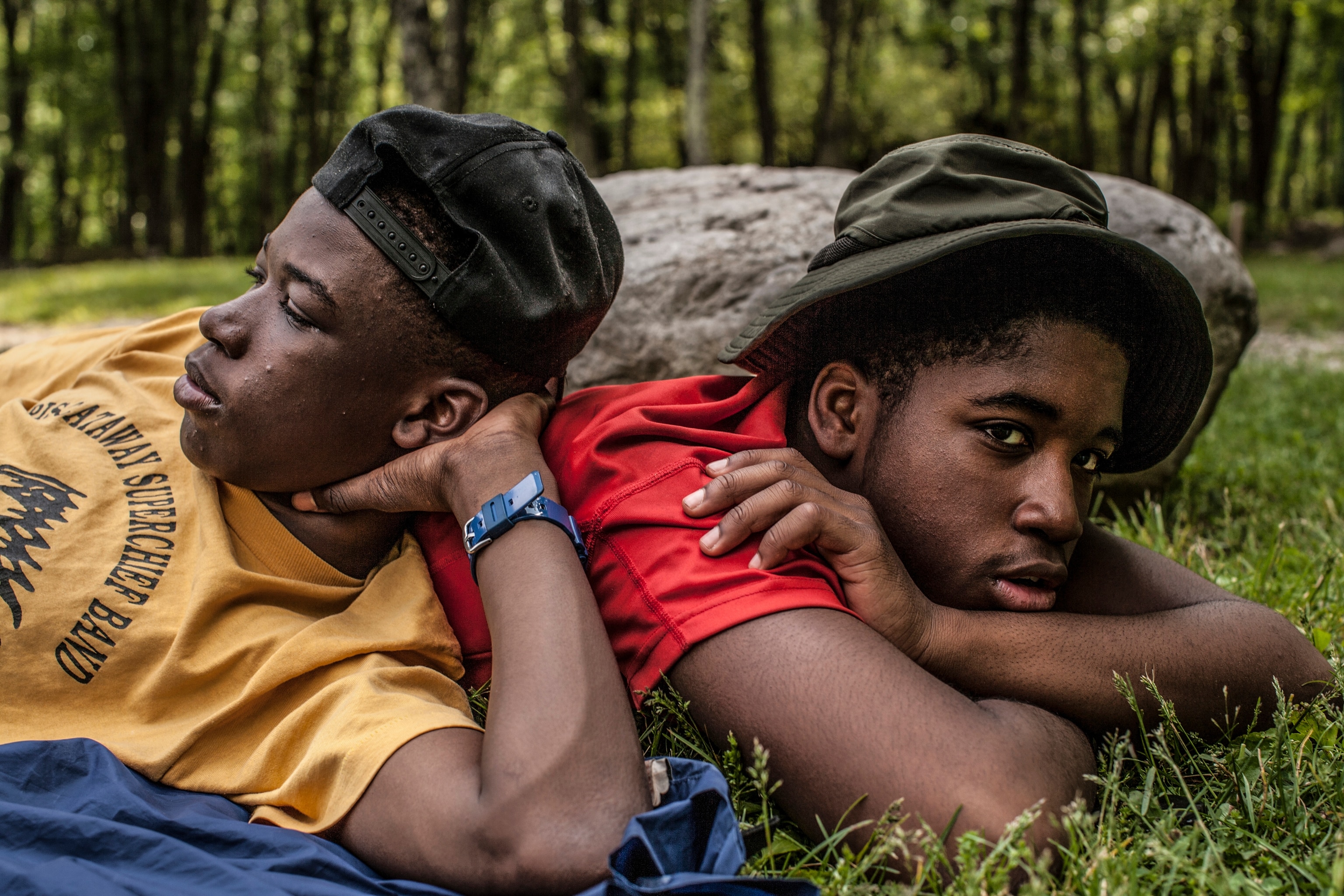 Two boys wearing hats lay down in the grass in a wooded area.