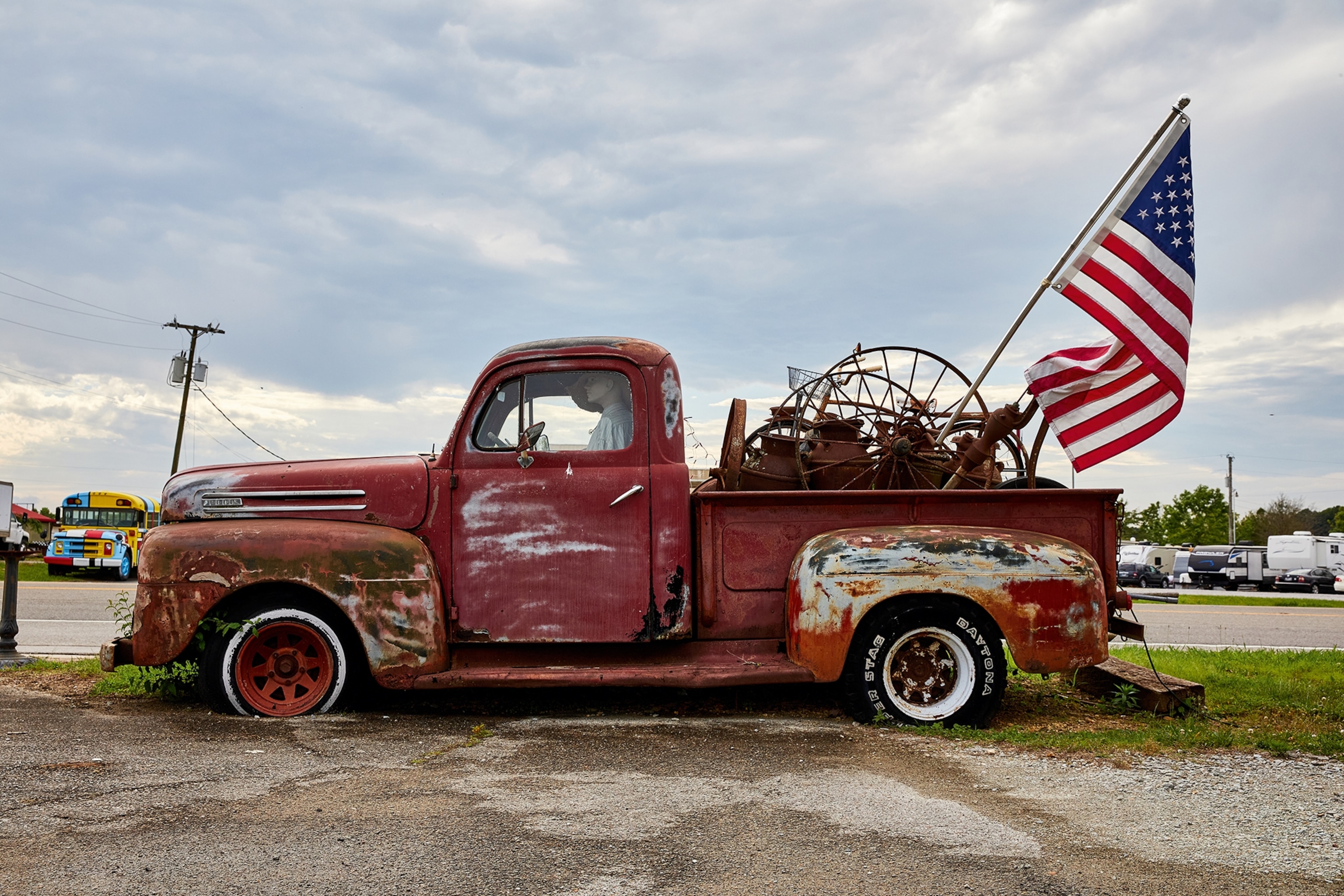 An imposing shot of a rusty and stalled pickup truck on the side of the road with an American flag waving from the back.