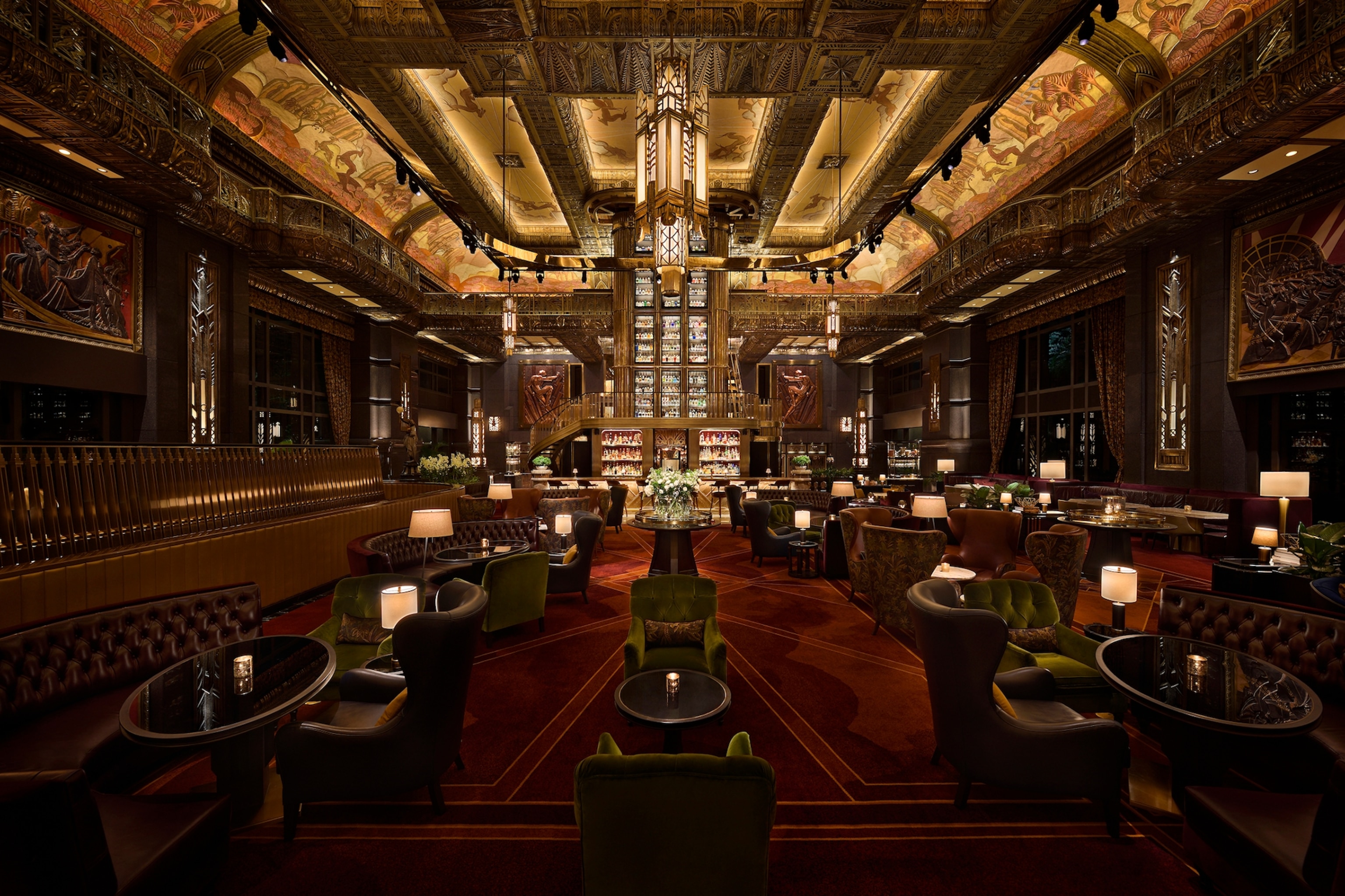 A grand room with a bar in the centre, surrounded by table lamps and velvet booths.