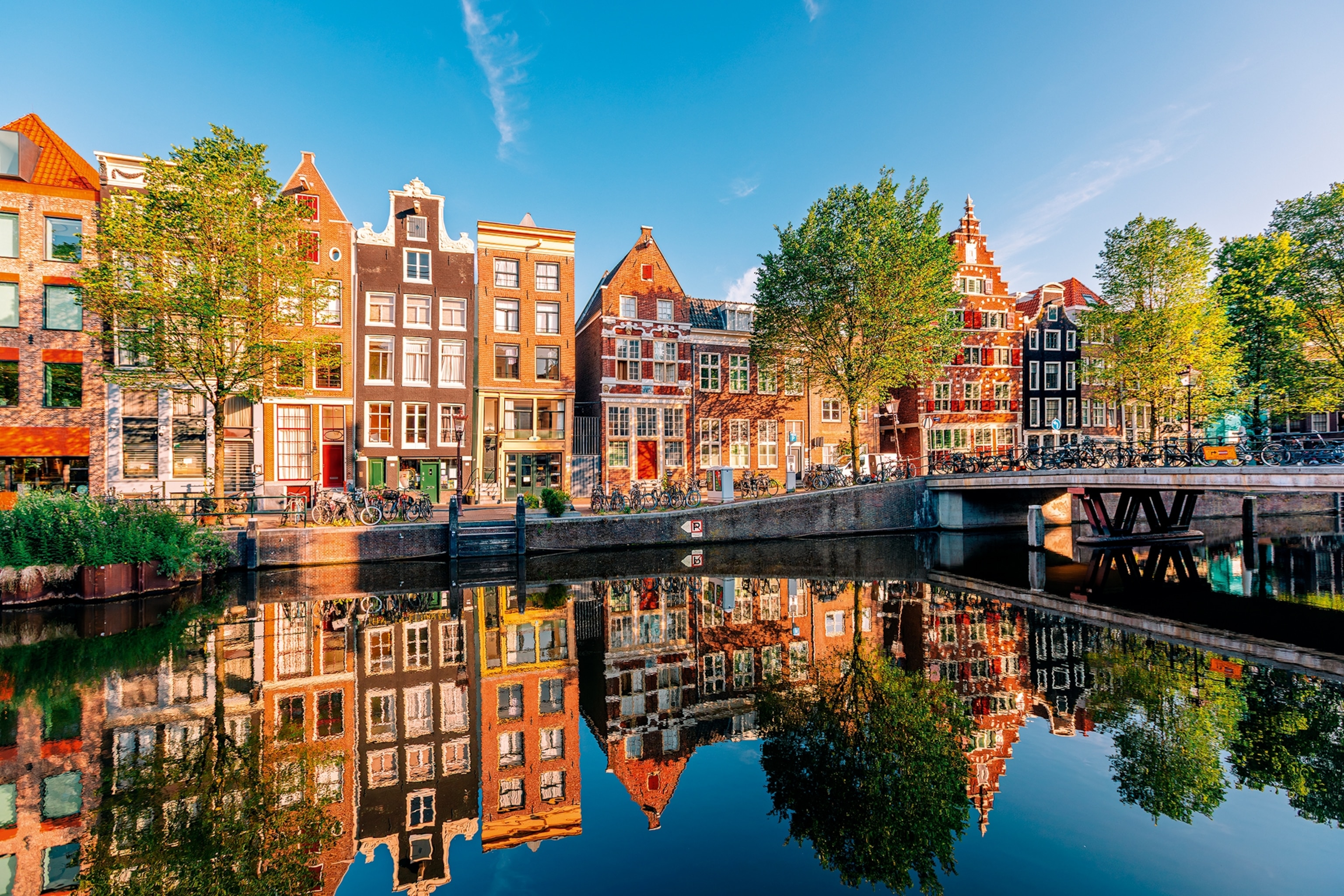 An urban canal scene in summer with Dutch flat buildings lining the water and trees marking either side of a bridge.