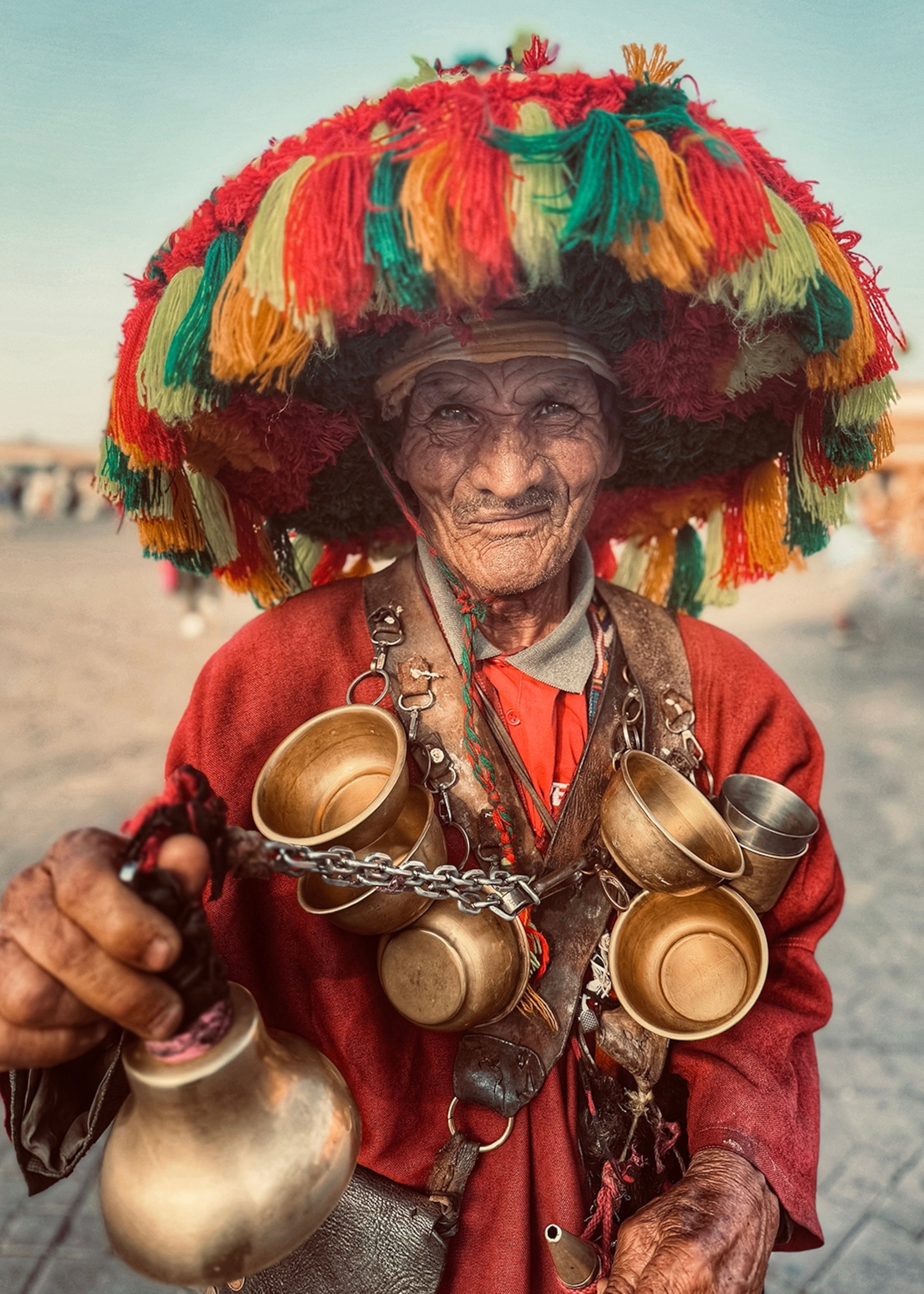 A portrait of an elderly water seller in Morocco wearing a tassel hat and tins cups on a chain around his neck.