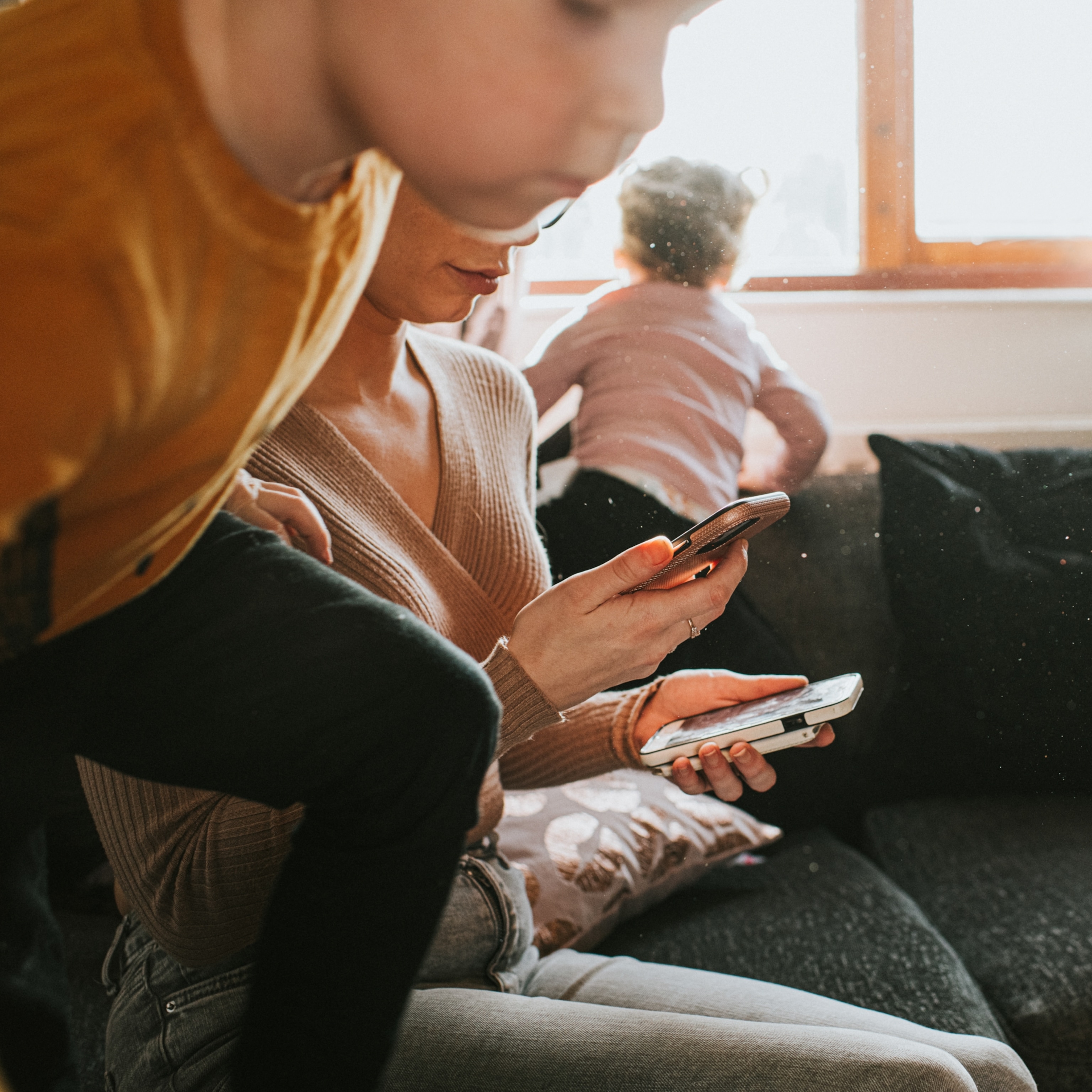 A woman sits on a couch, intently looking at two smartphones. A child in a yellow shirt climbs near her, with another child at the window, creating a busy, distracted atmosphere.