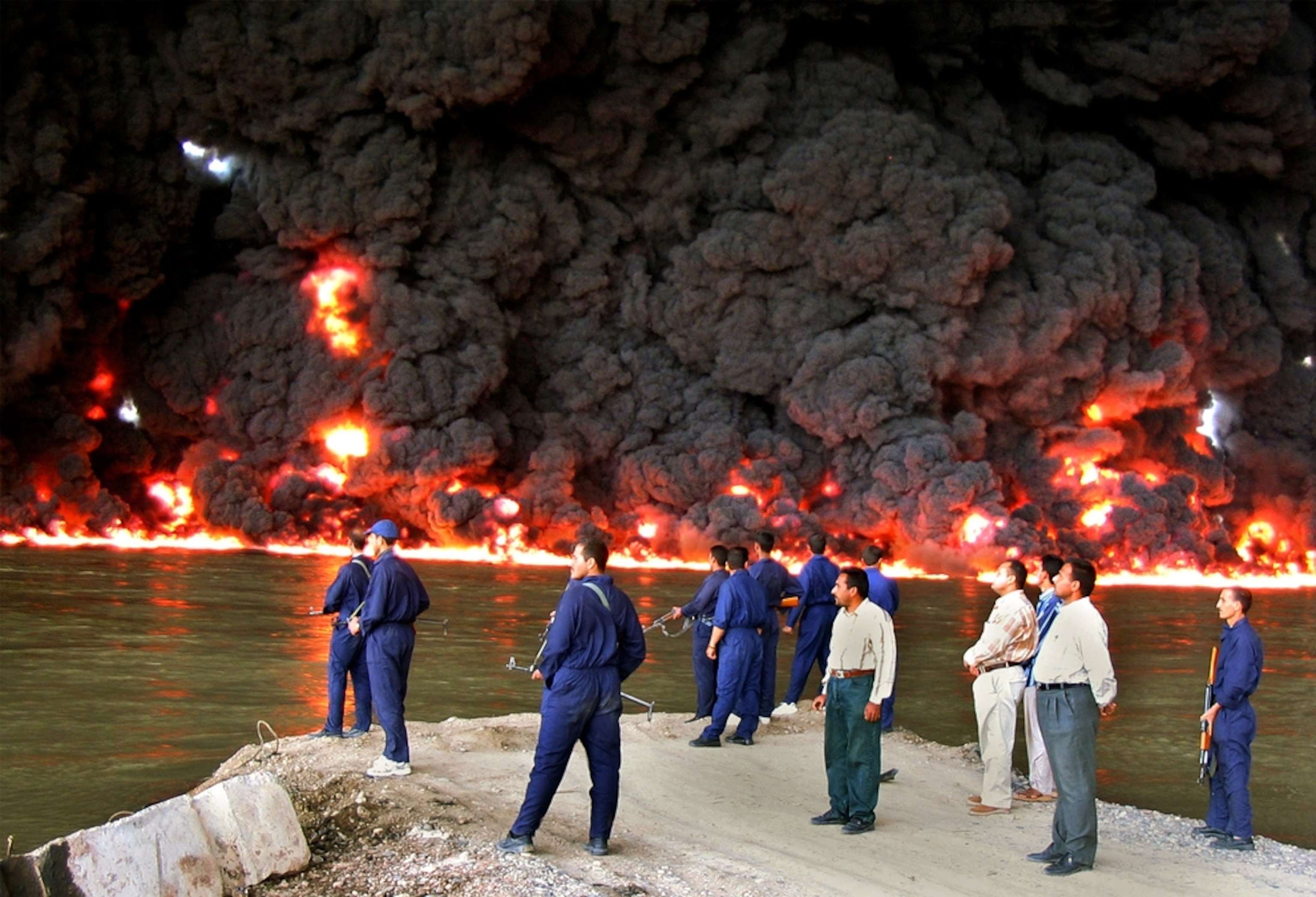 Iraqi police officers watch fire and smoke billow from a pipeline fire.