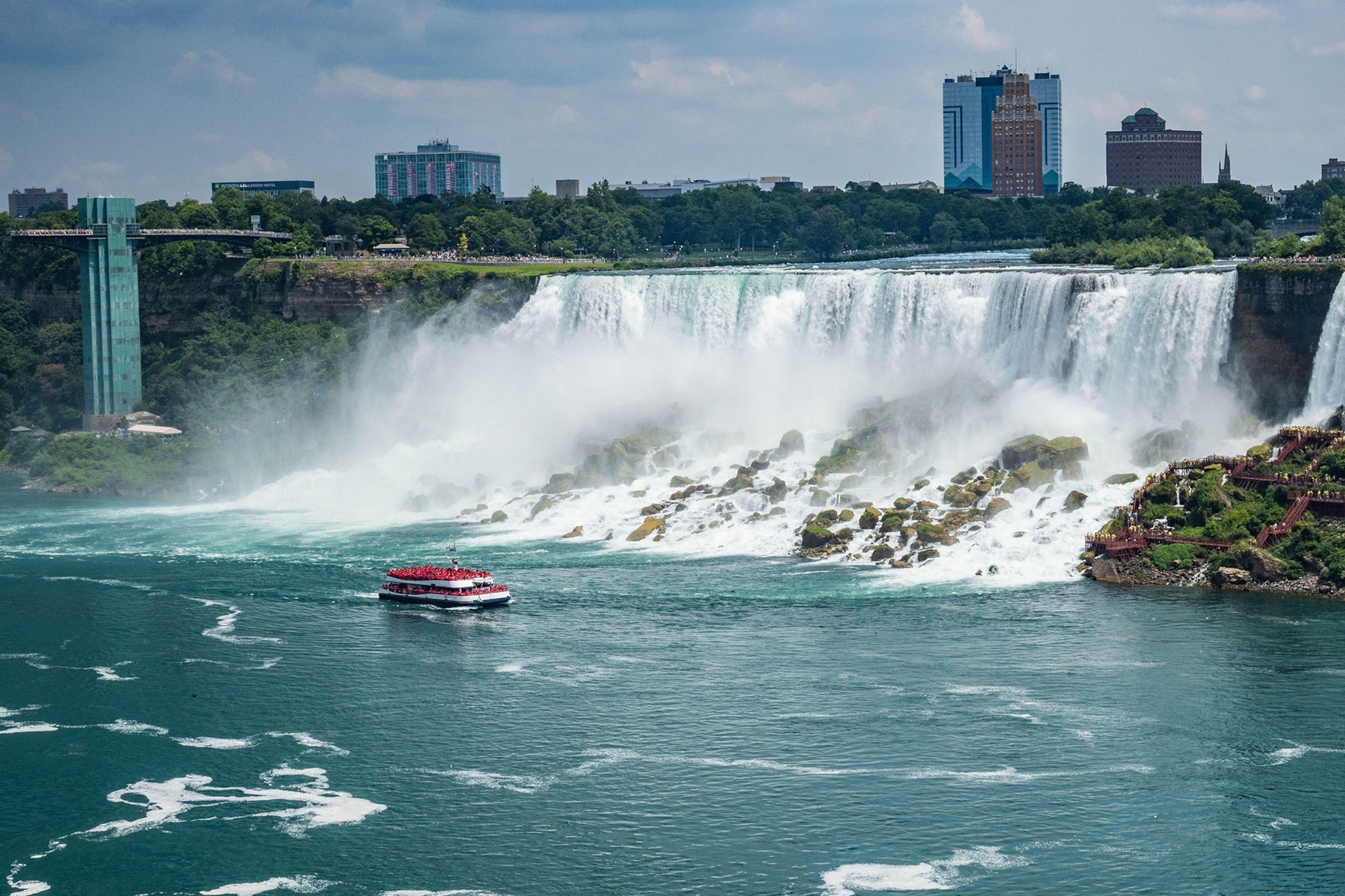 A tourist boat floats past a huge waterfall with city buildings in the background.