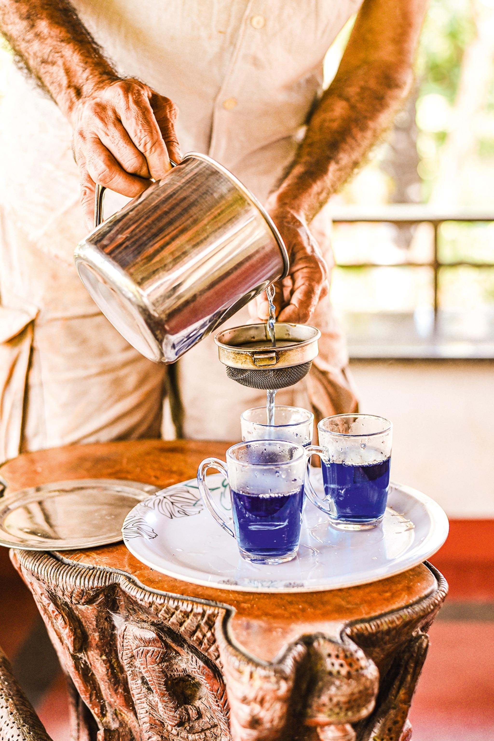 A man pouring a uniquely blue-coloured tea through a sieve into small glass cups on a serving plate.