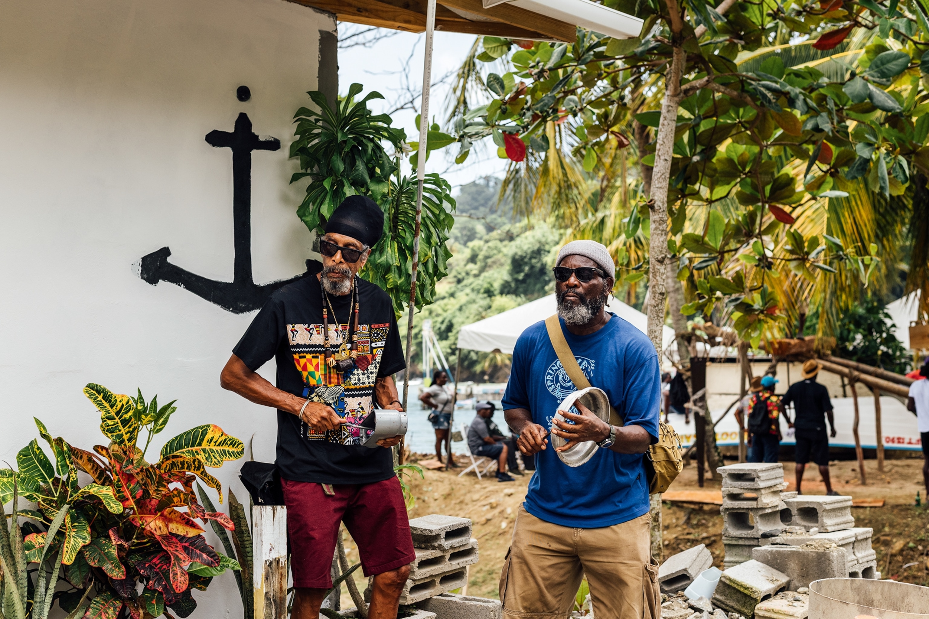 Two relaxed men with sunglasses playing makeshift instruments in front of a simple house with tents in the back.