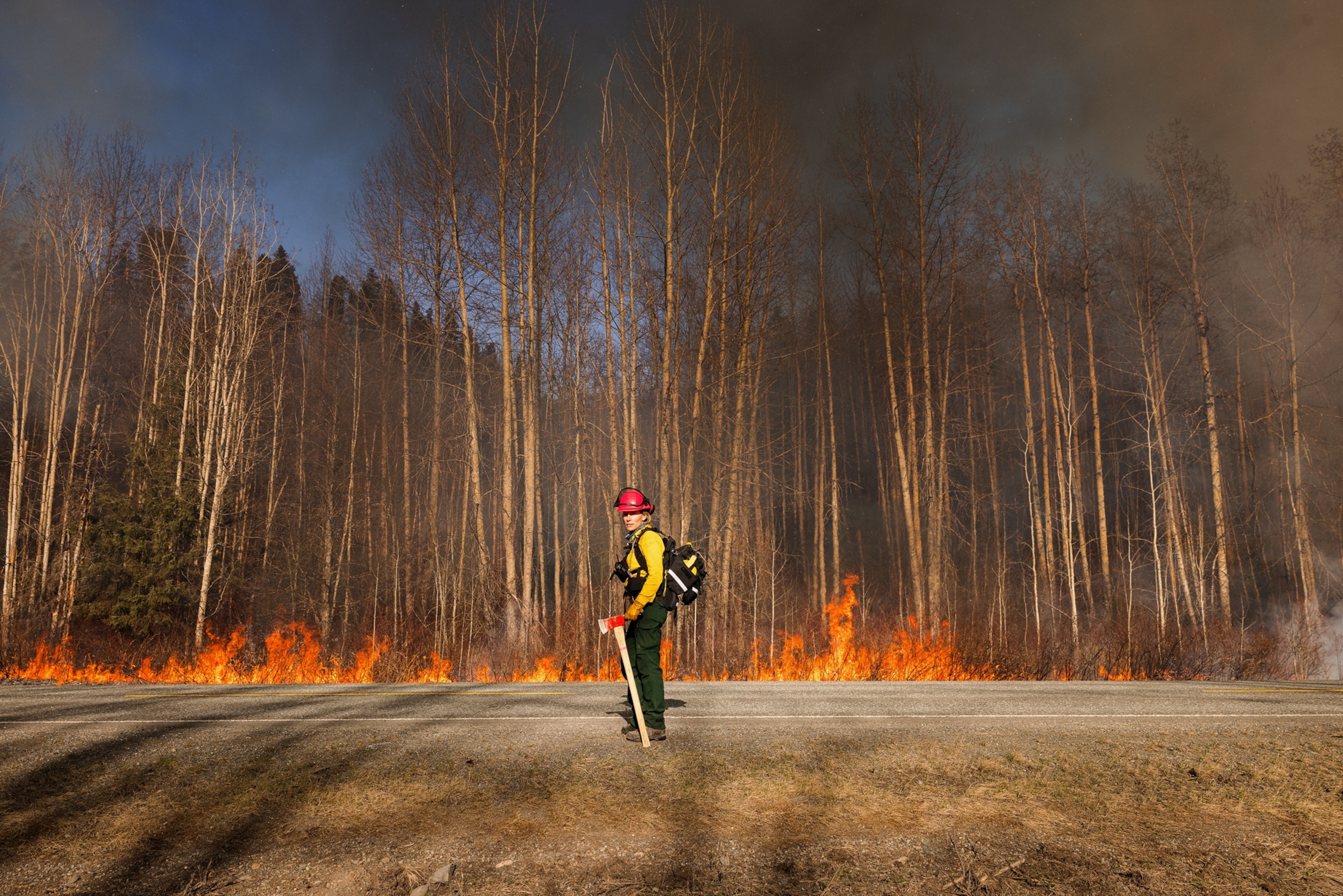 A woman stands in front of a line of trees that begin to burn