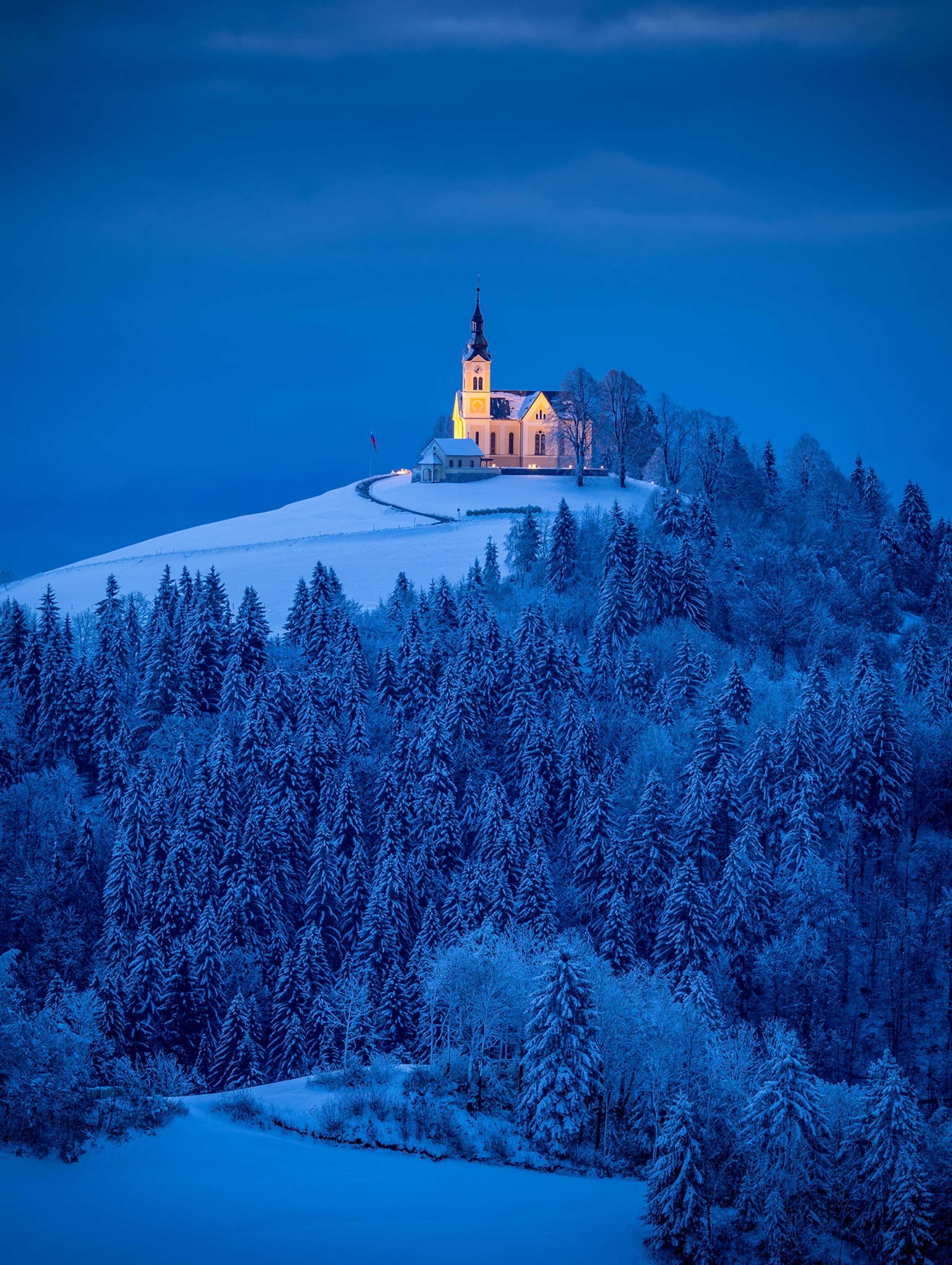 In the early morning blue hour, St. Lenart Church in Crni Vrh, Slovenia, stands like a beacon above the snow-covered landscape.