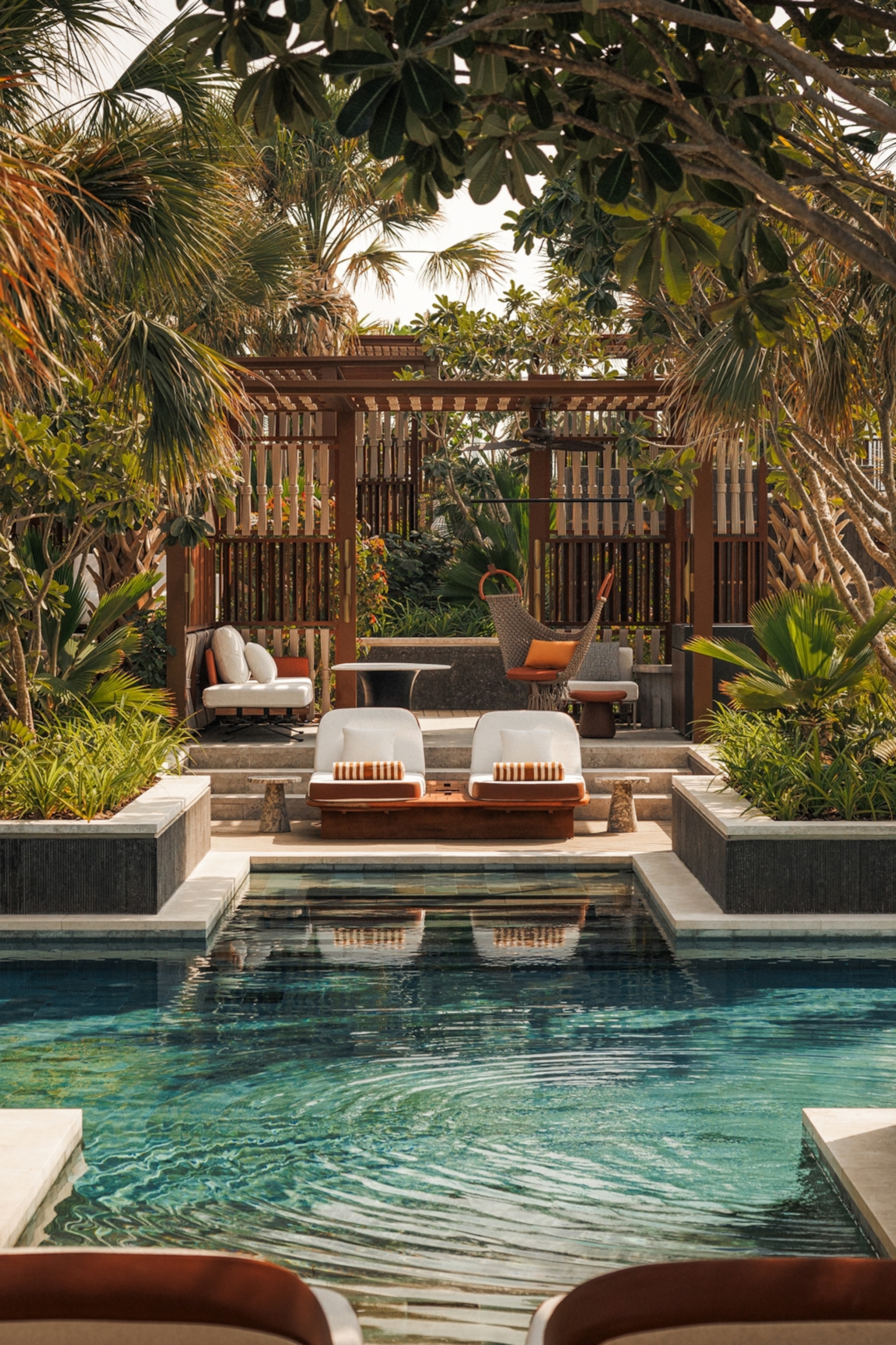 A symmetrical pool in the shape of a cross with two lounge chairs on one end and trees bending over above.