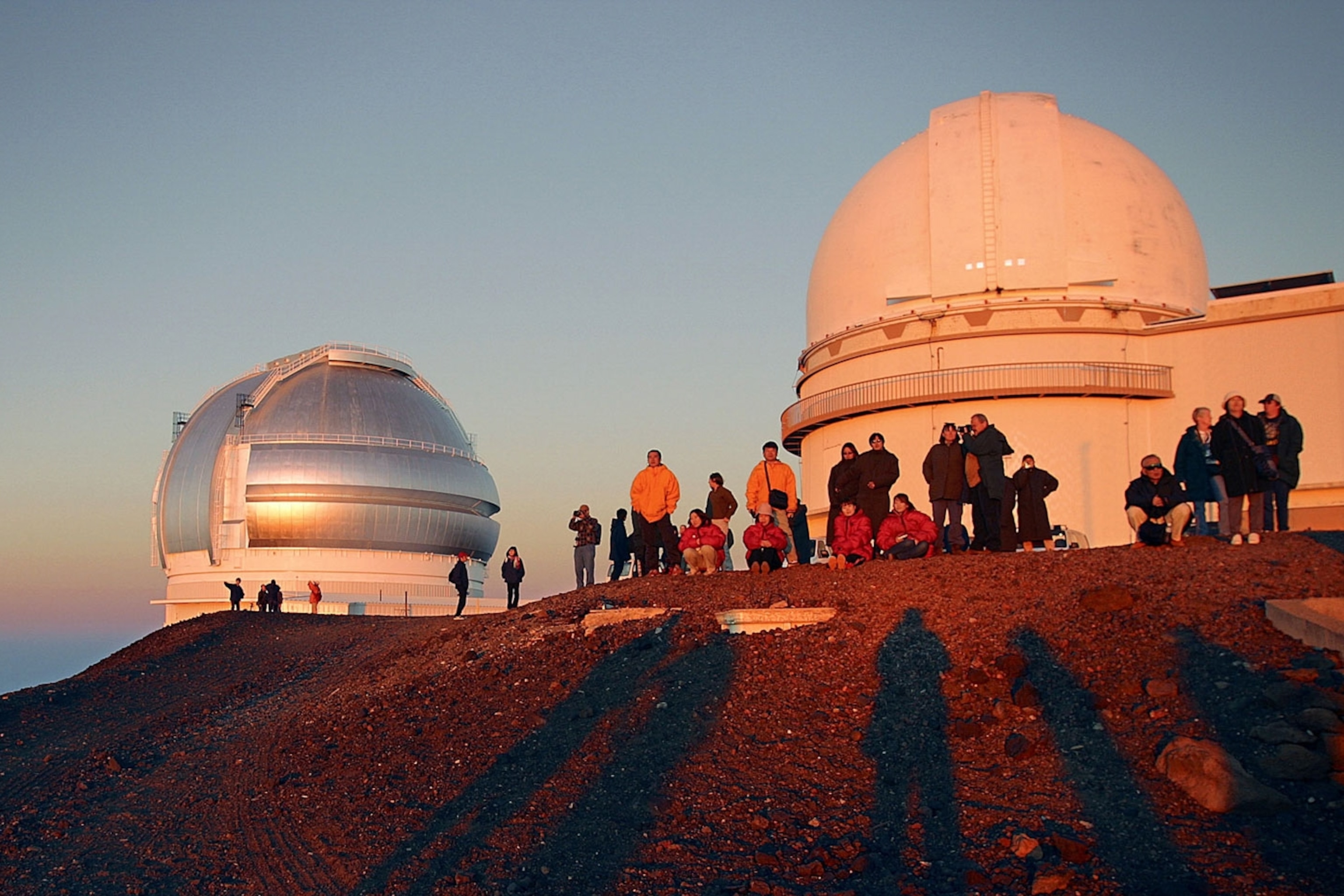 A group of people gather at sunset on a rocky hilltop in front of two telescopes.