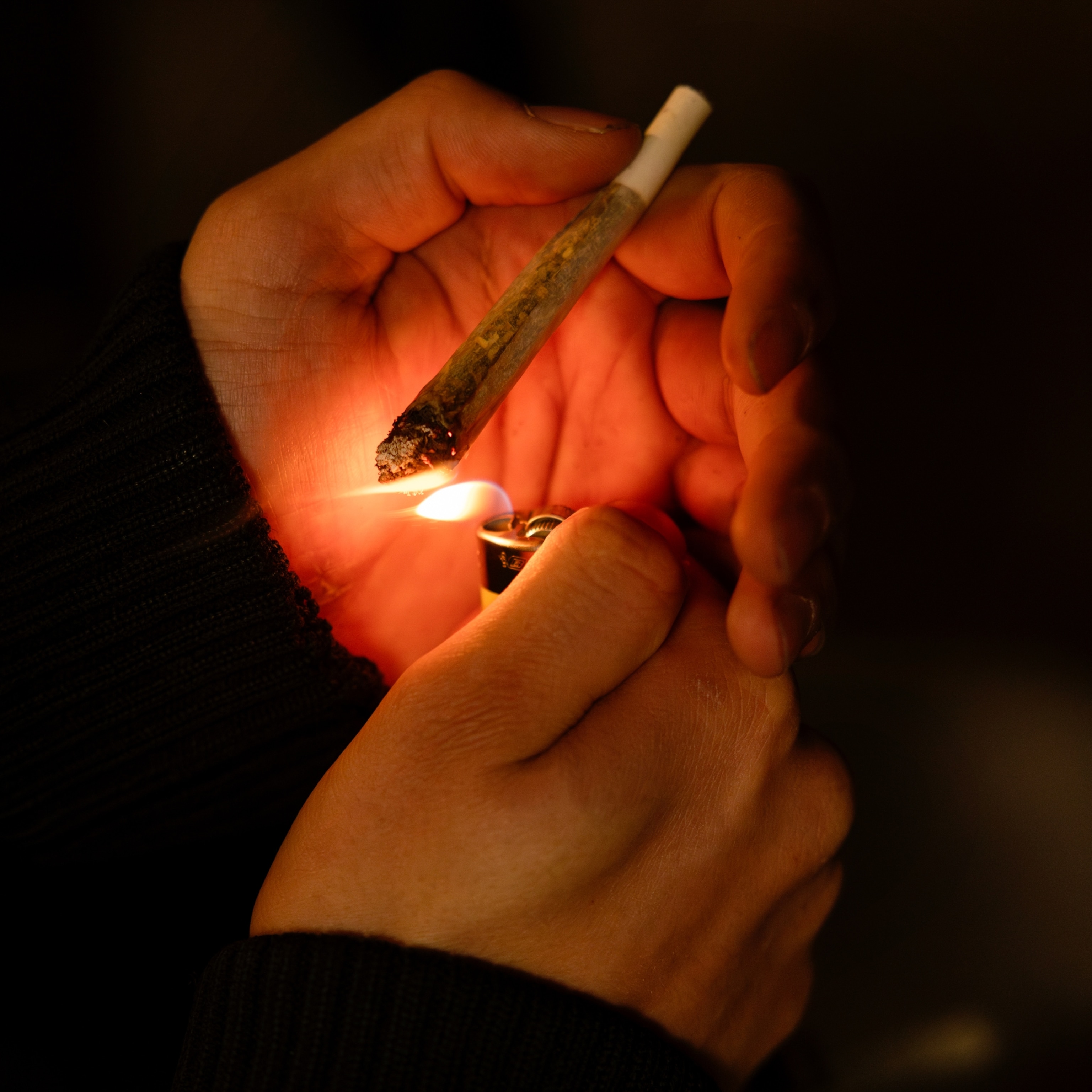Hands hold a marijuana joint and lighter, illuminating the scene with a warm glow against a dark background