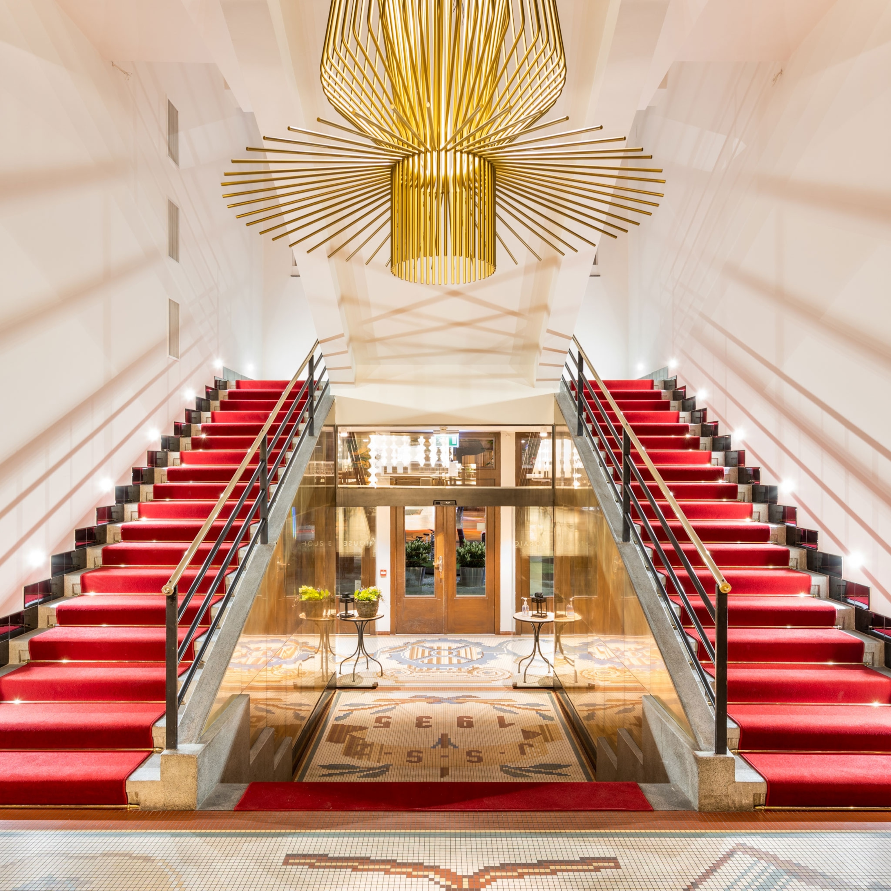 Elegant interior with twin red-carpeted staircases rising on either side, a large gold chandelier overhead, and mosaic tile flooring leading to glass double doors.
