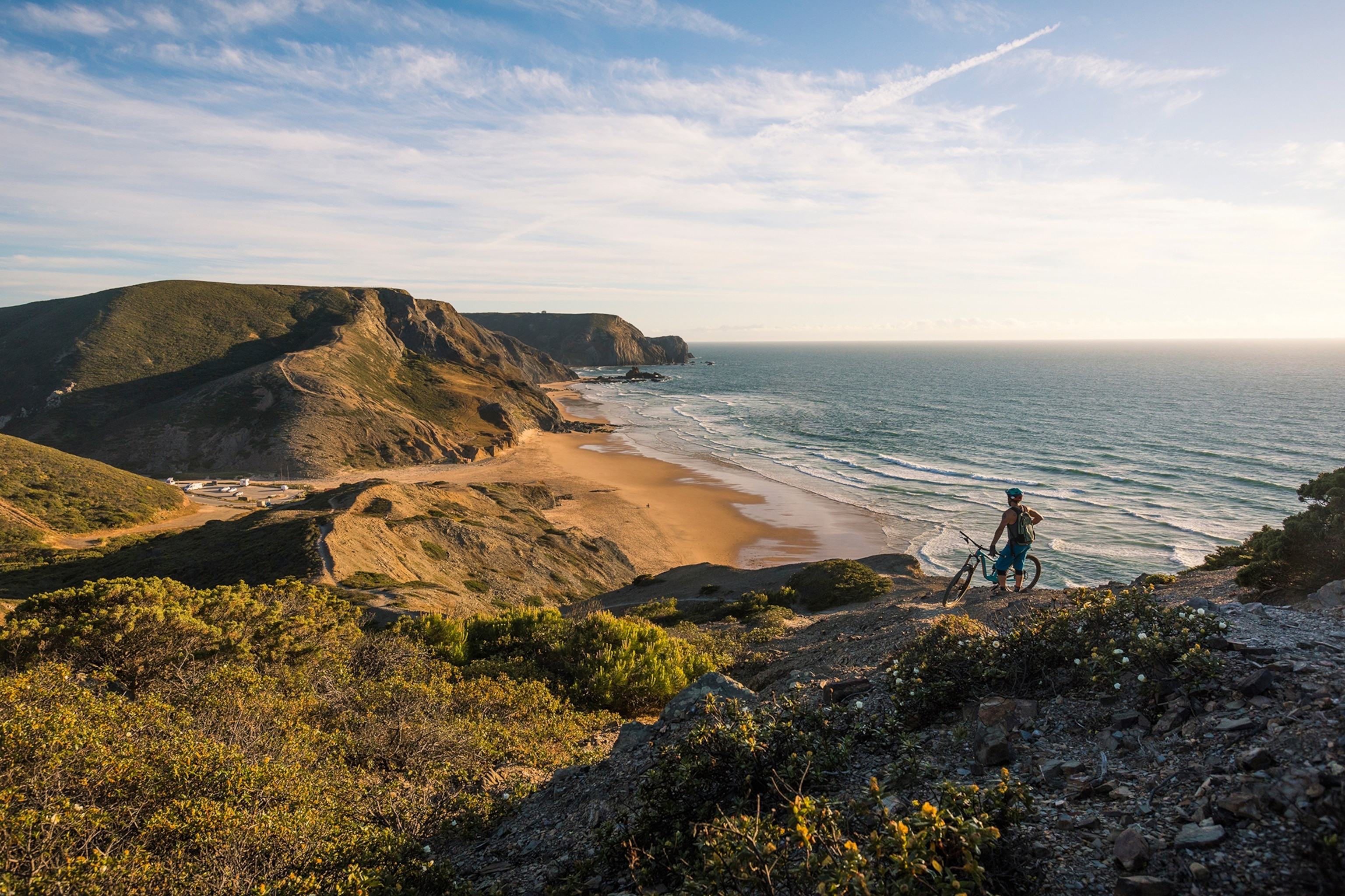 A person with their bike standing on the edge of a low cliff, overlooking a windy beach wit low waves.