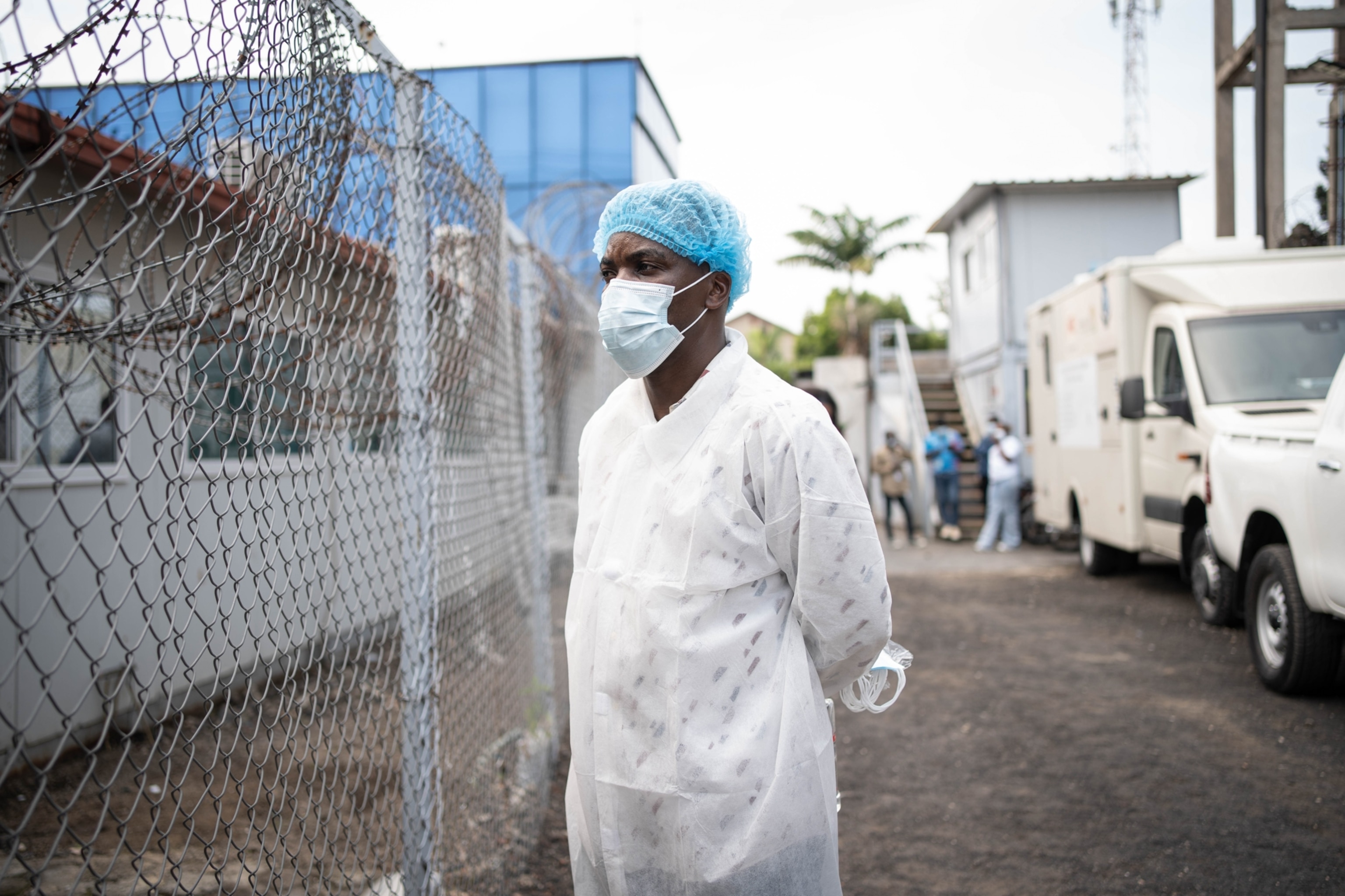 A healthcare worker in protective gear, including a mask and hairnet, stands near a chain-link fence by a medial field facility